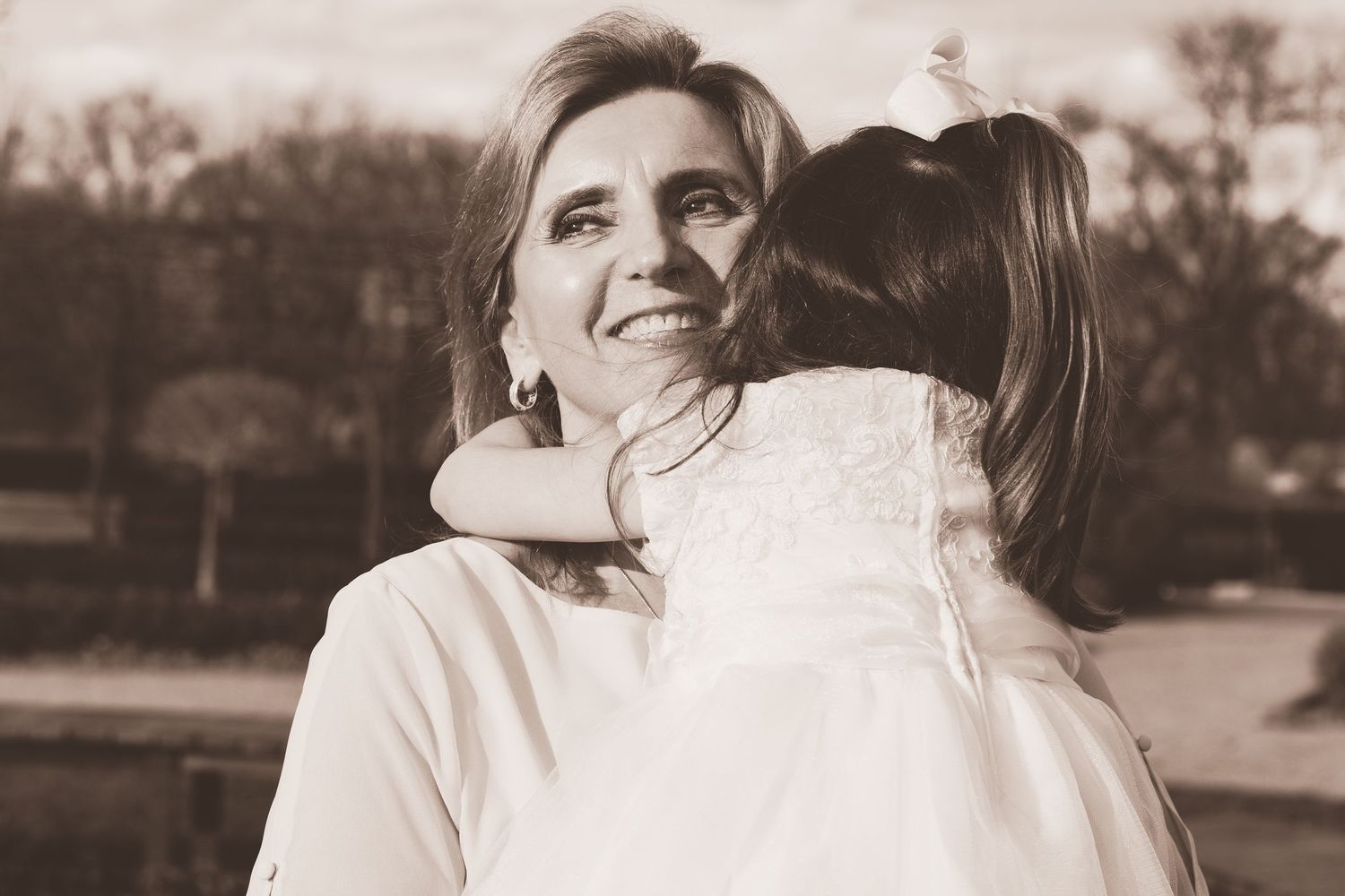 Sepia toned photo of smiling woman embracing young girl in white dress outdoors.