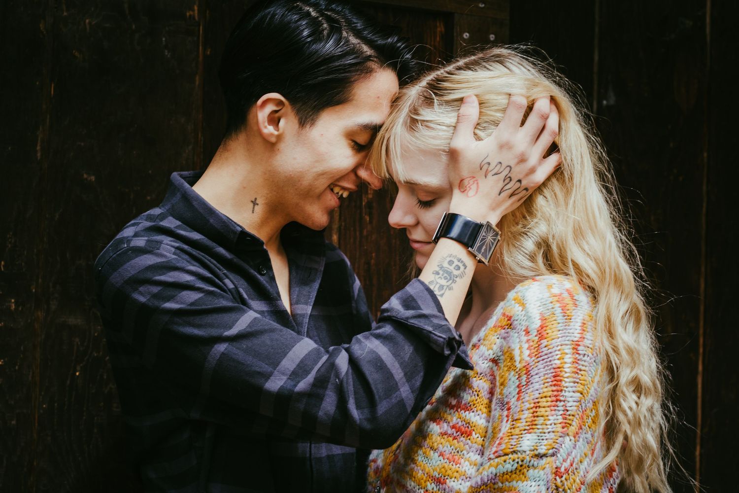 Two people sharing an intimate moment, touching foreheads tenderly, one with tattoos and blonde hair, outdoors.