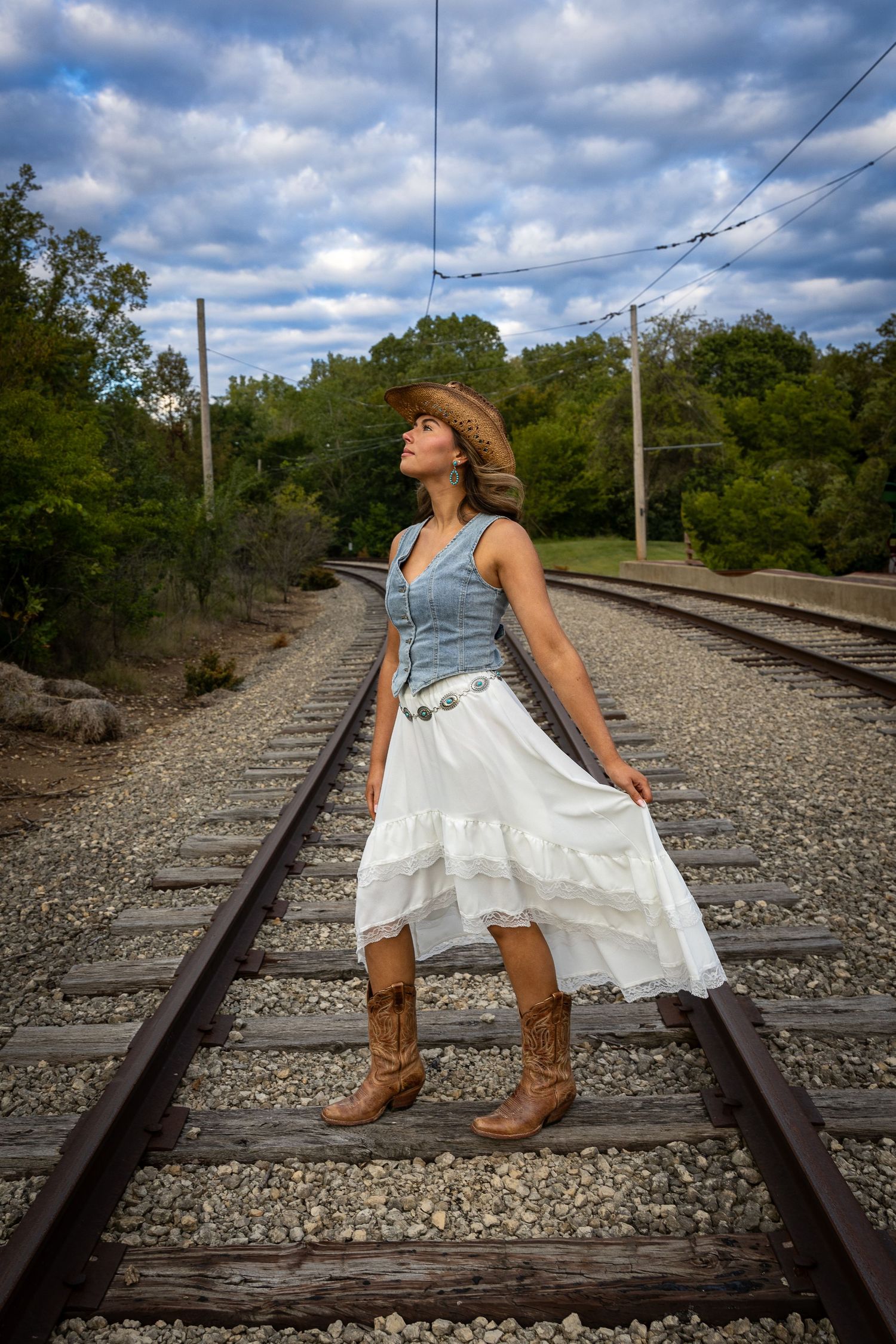 Woman in cowboy hat, white skirt, and boots standing on railroad tracks looking up at cloudy sky.