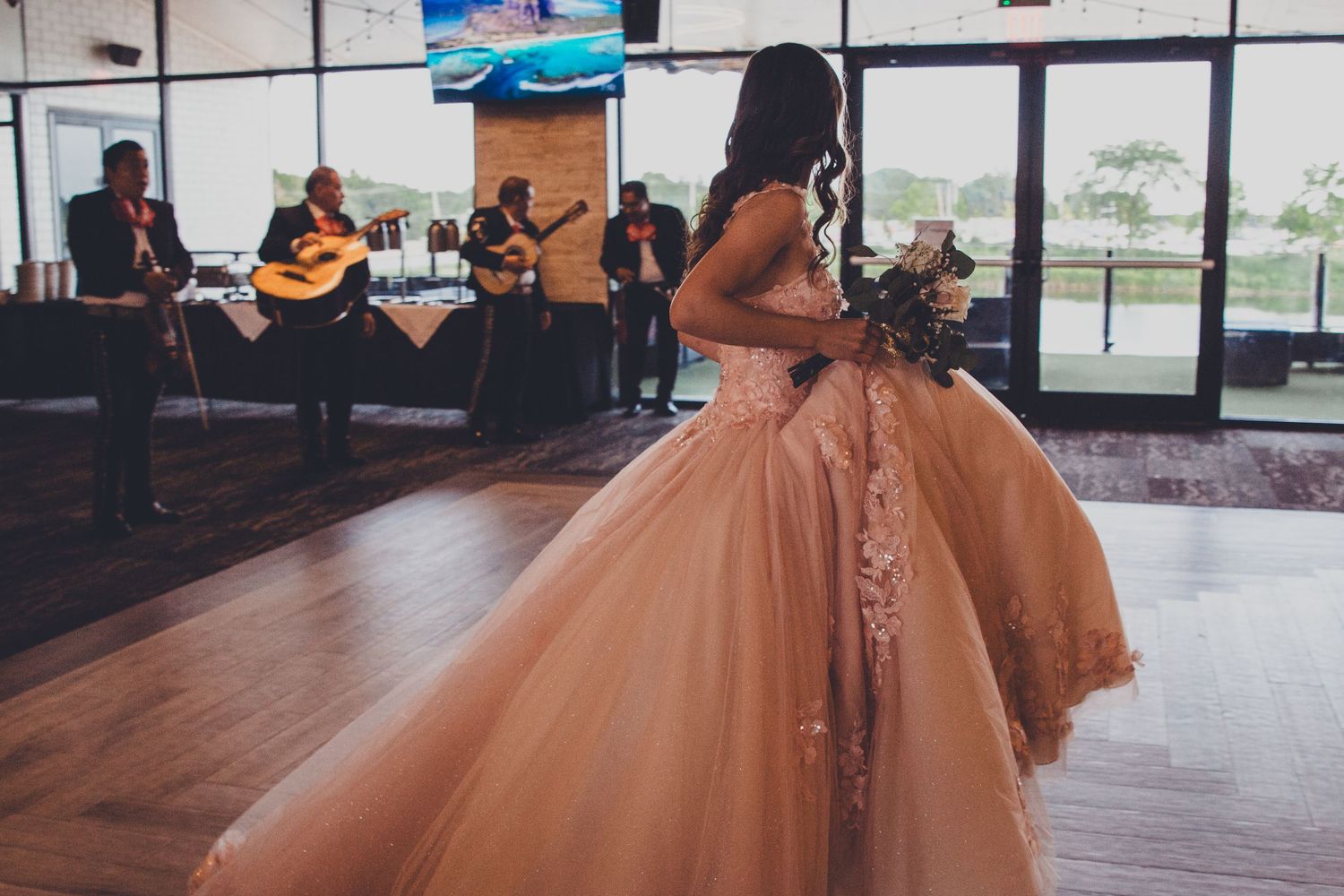 Young woman in blush ball gown dances as a mariachi band performs on stage at her quinceañera celebration.
