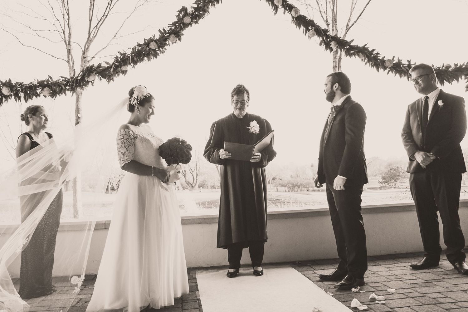Black and white wedding ceremony photo with bride, groom, and officiant at an outdoor altar.