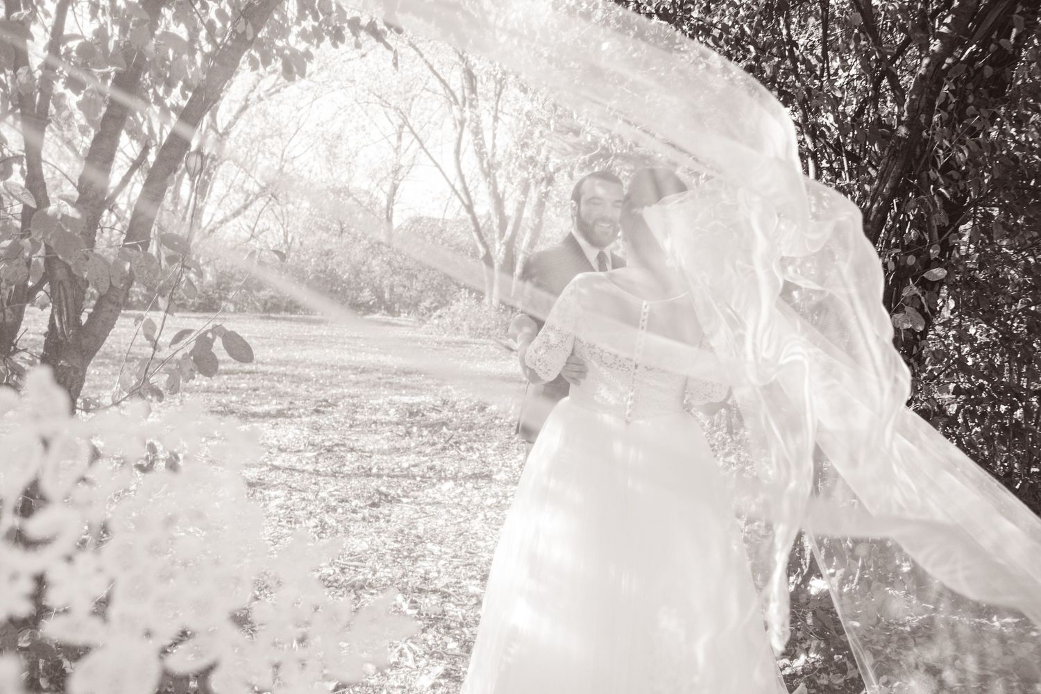 Black and white photo of bride and groom embracing outdoors with flowing wedding veil blowing in the wind.