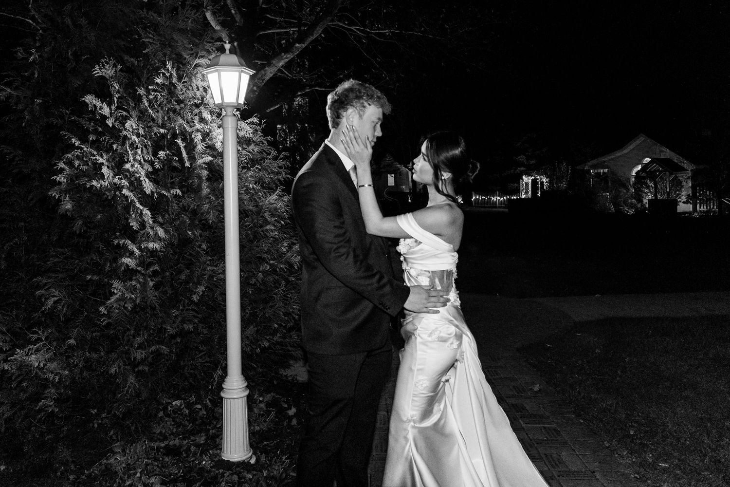 Black and white photo of a bride and groom sharing a romantic kiss under a glowing street lamp at night.