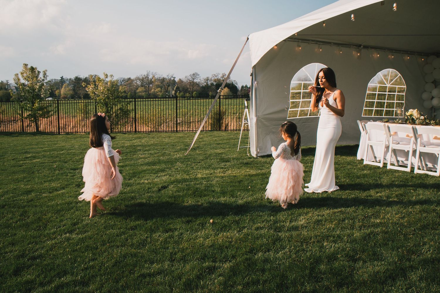 Bride in white gown greets two flower girls in pink tutus on green lawn near wedding tent.