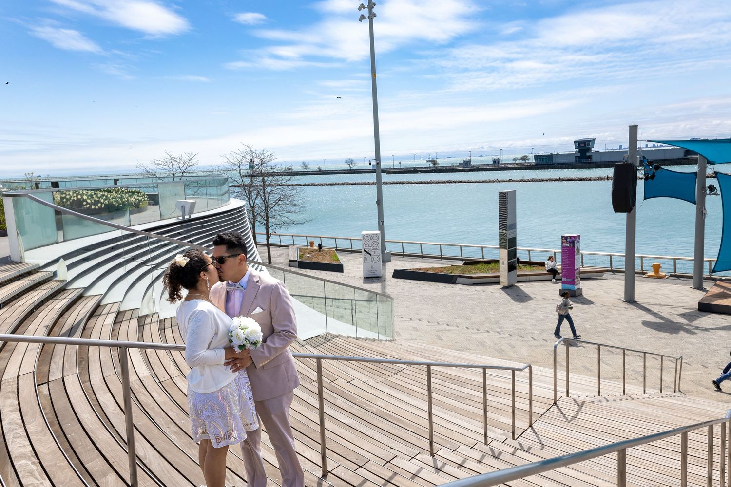 Newlywed couple sharing a tender kiss on a waterfront boardwalk under a bright blue sky.