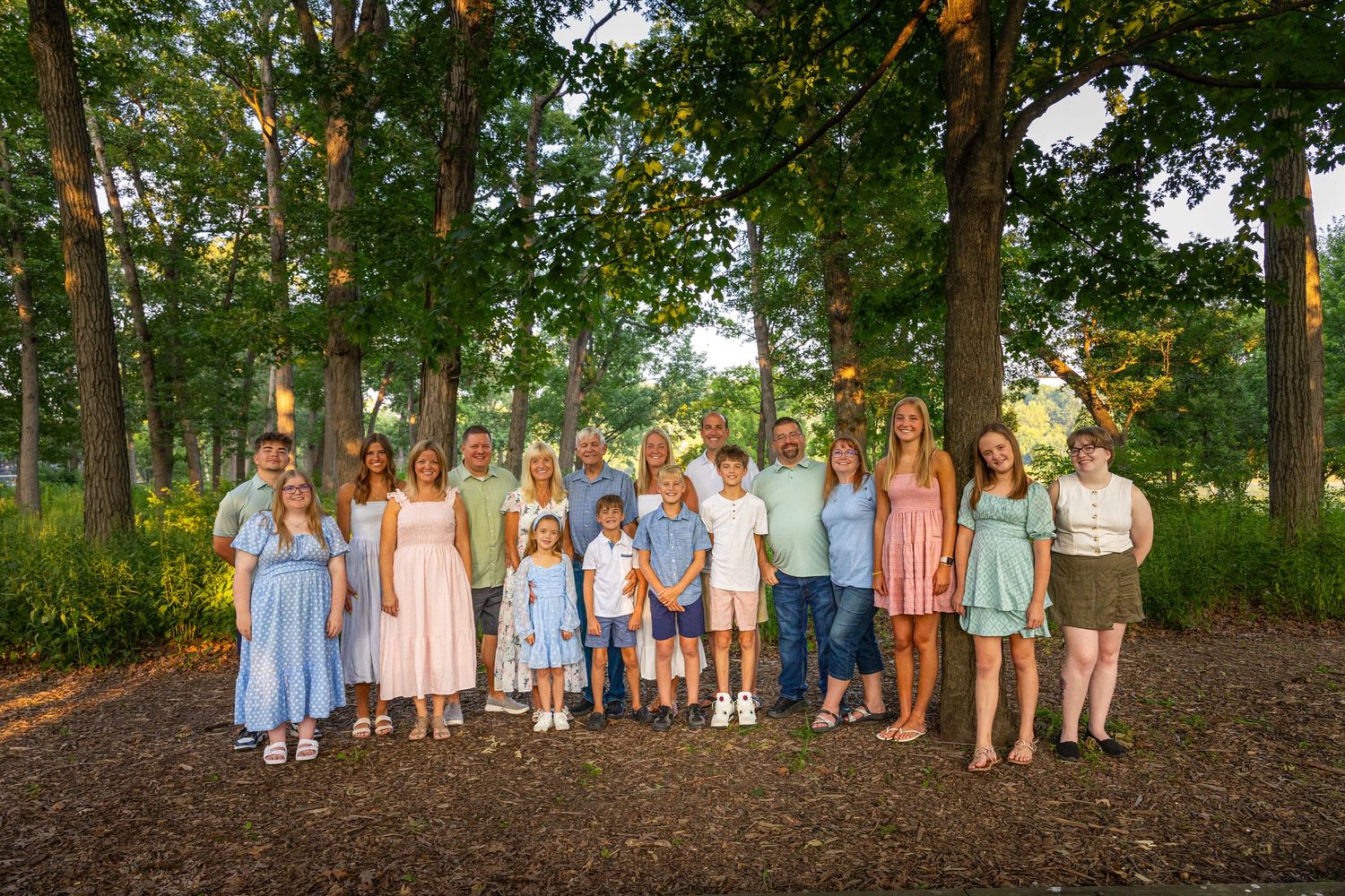 Large extended family portrait taken outdoors among tall trees during golden hour.