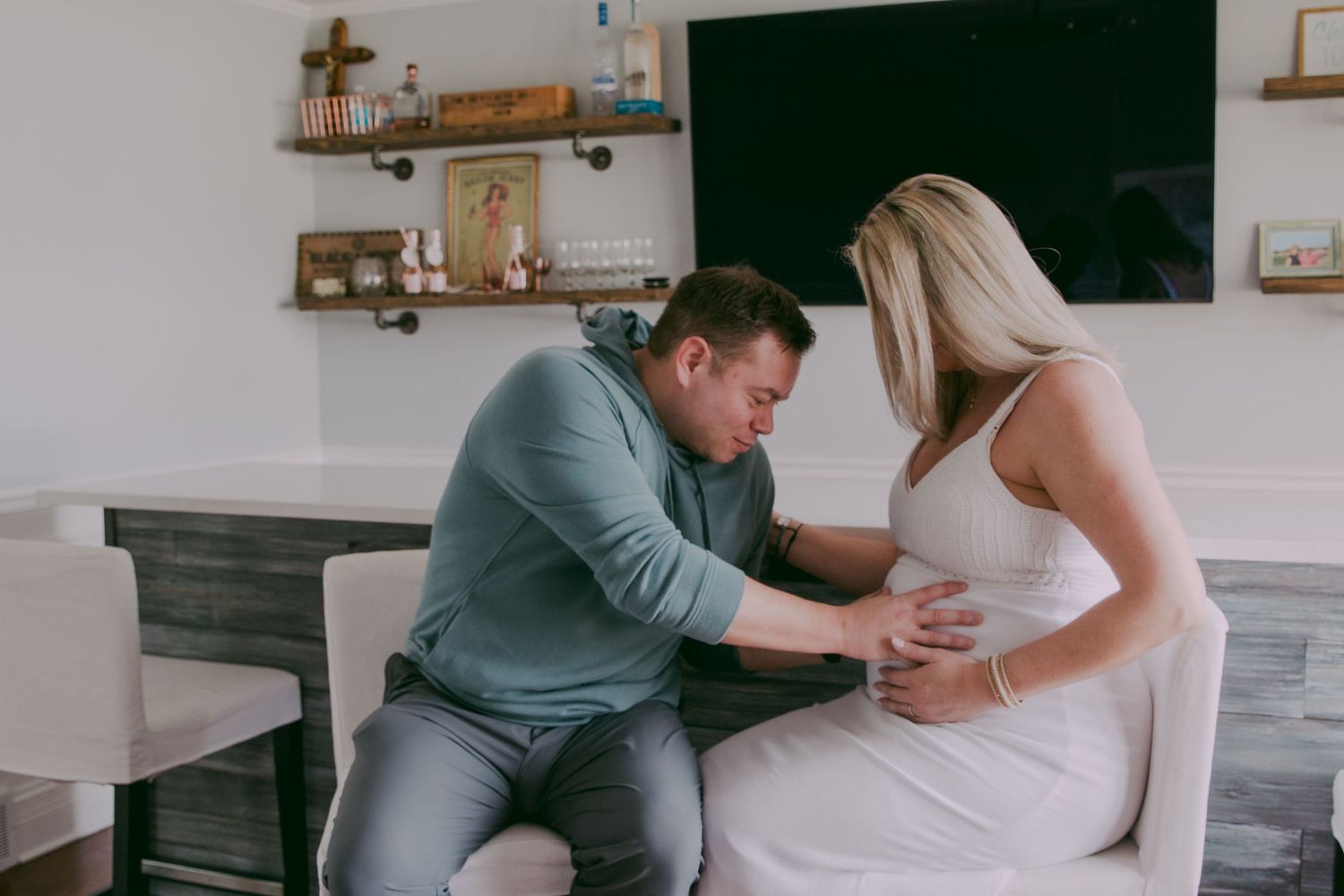 Tender moment as man leans to listen to pregnant partners baby bump indoors at home.