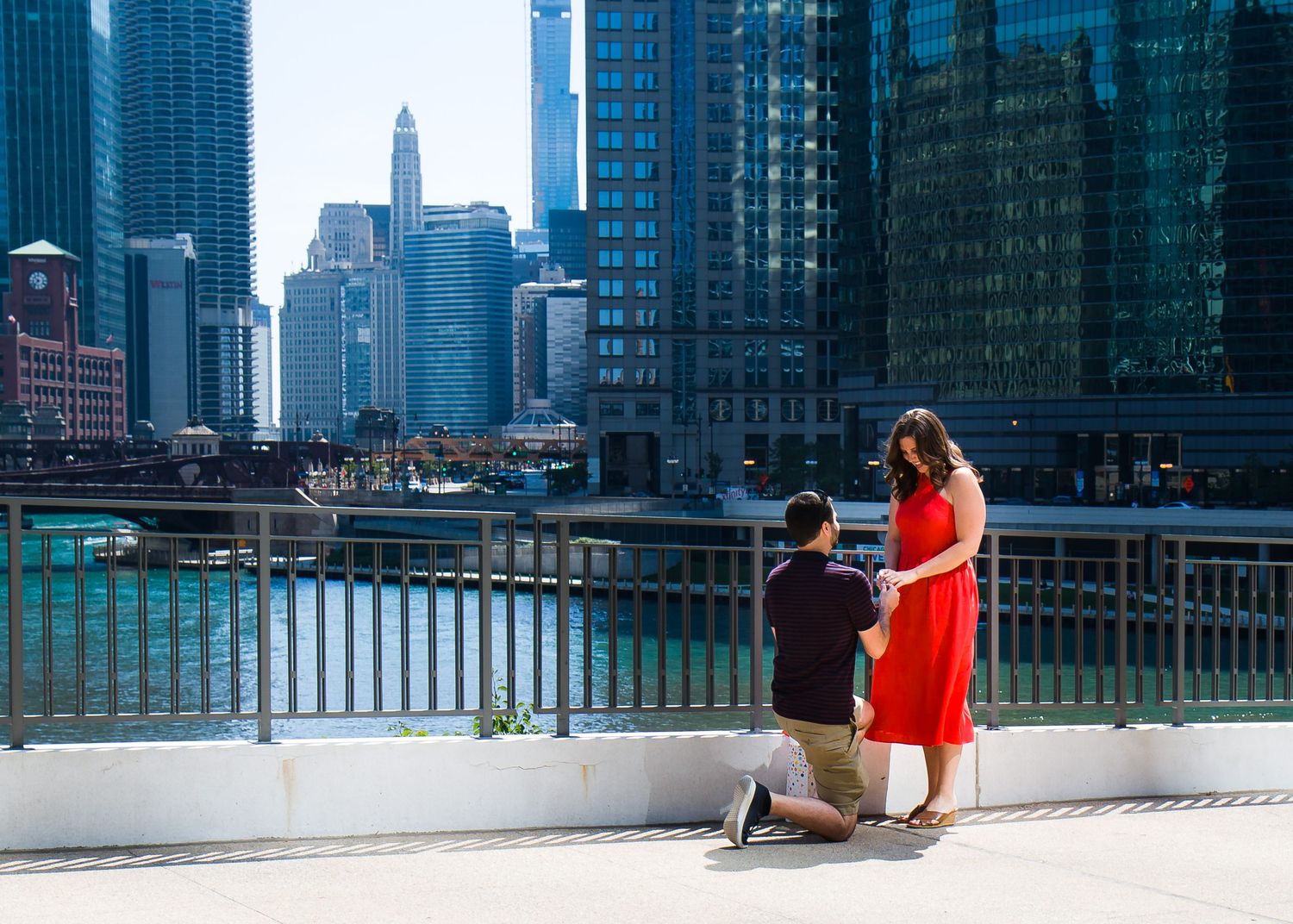 Man proposing on one knee to woman in red dress on Chicago Riverwalk with city skyline backdrop.