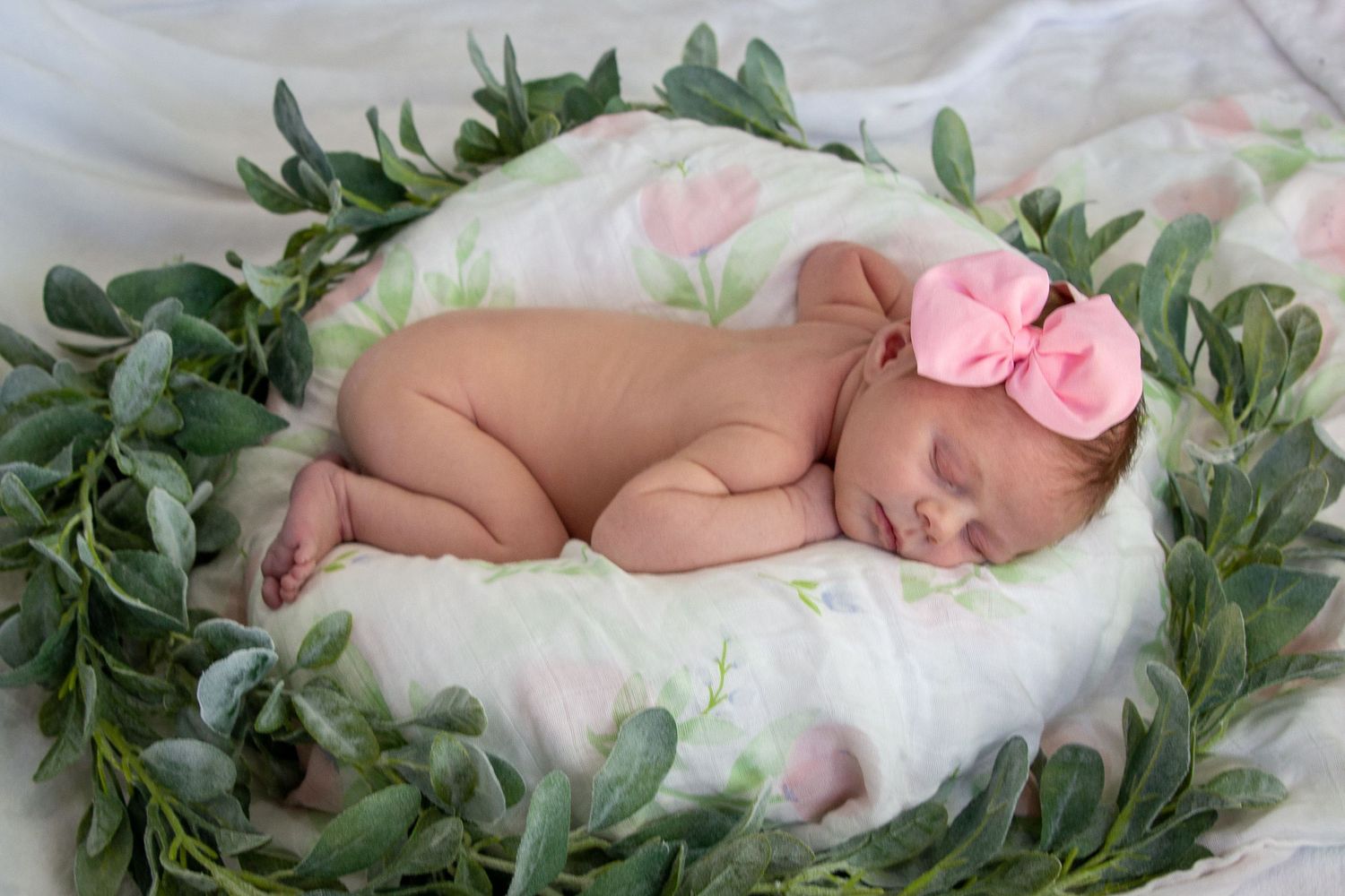 Sleeping newborn baby girl with pink bow surrounded by a wreath of green leaves.