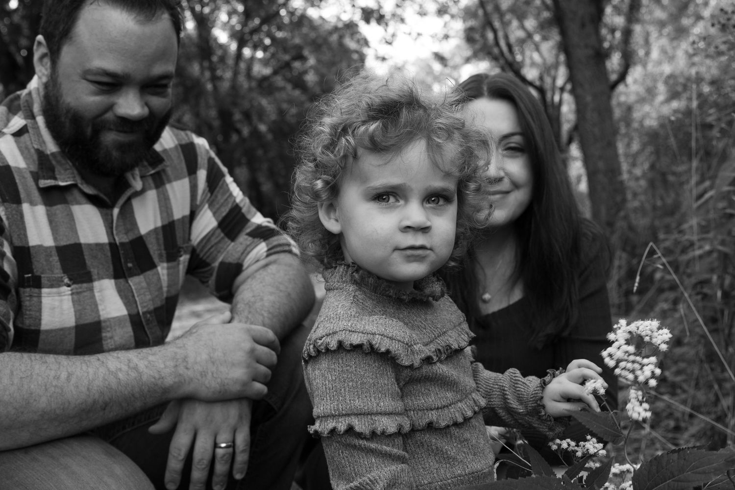 Black and white portrait of a couple with their young curly-haired toddler in a woodland setting.