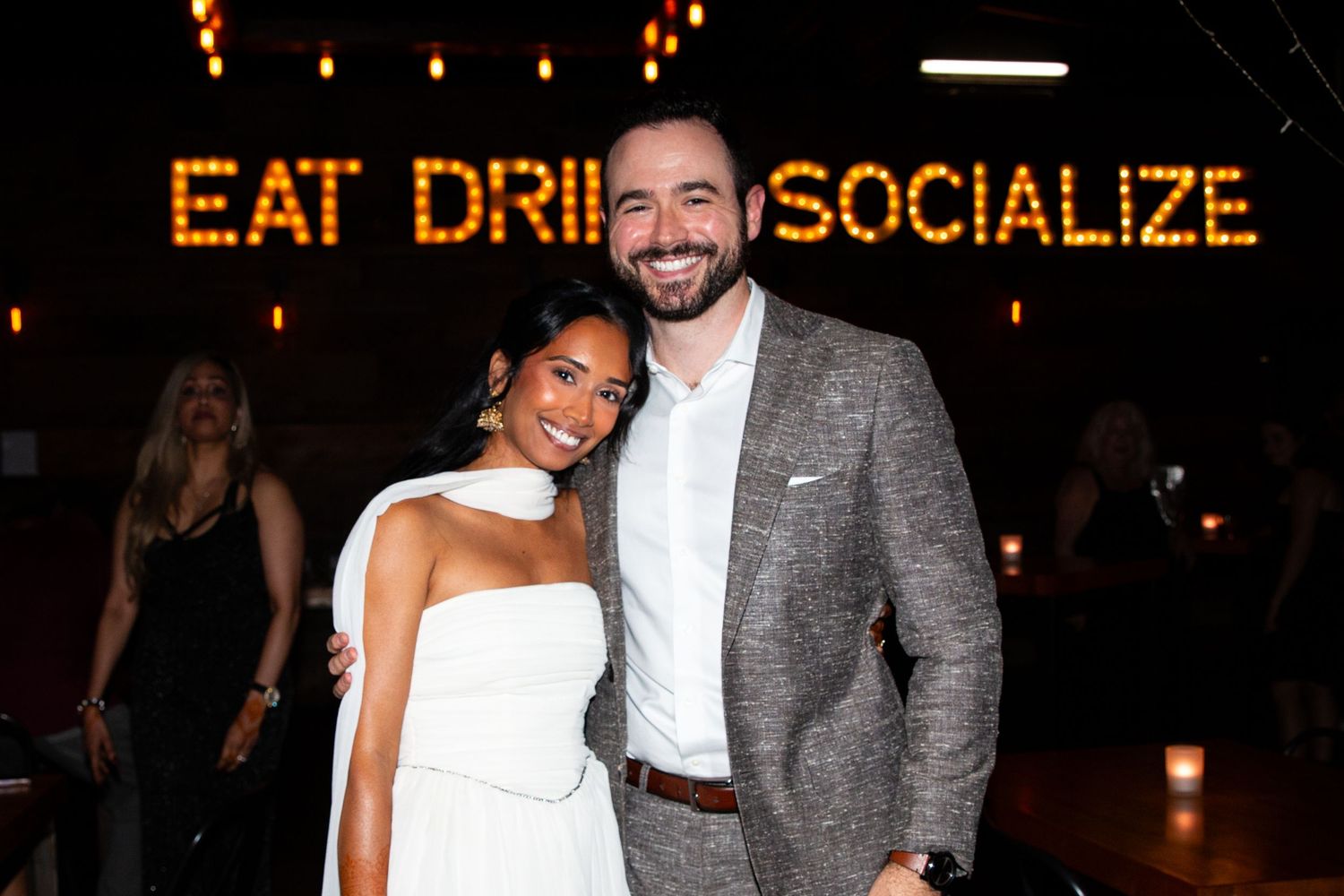 Smiling couple poses together at social event in front of lit marquee sign at venue.