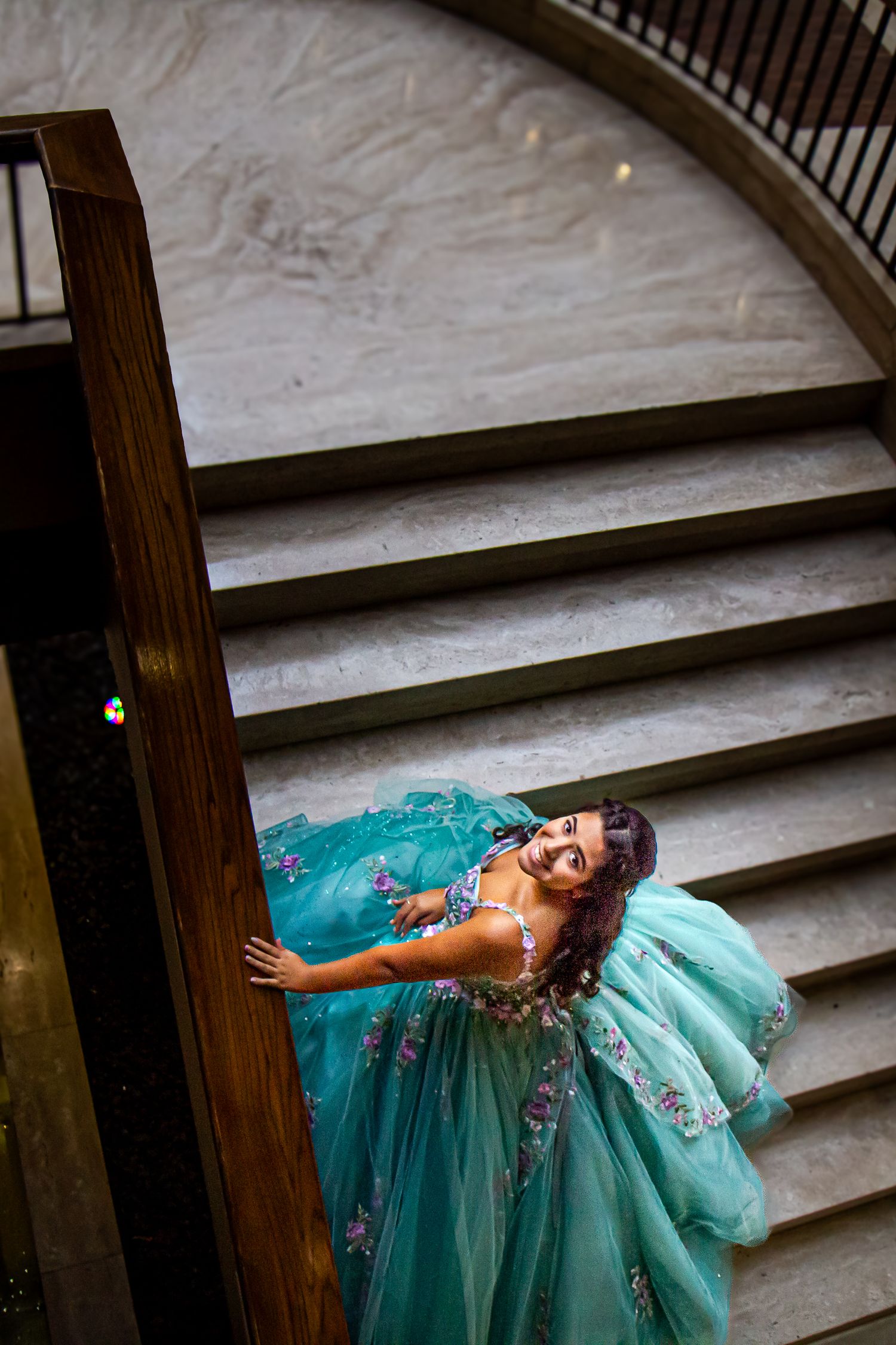 Young woman in teal floral ball gown poses on elegant curved staircase, smiling upward in a stunning quinceañera portrait.
