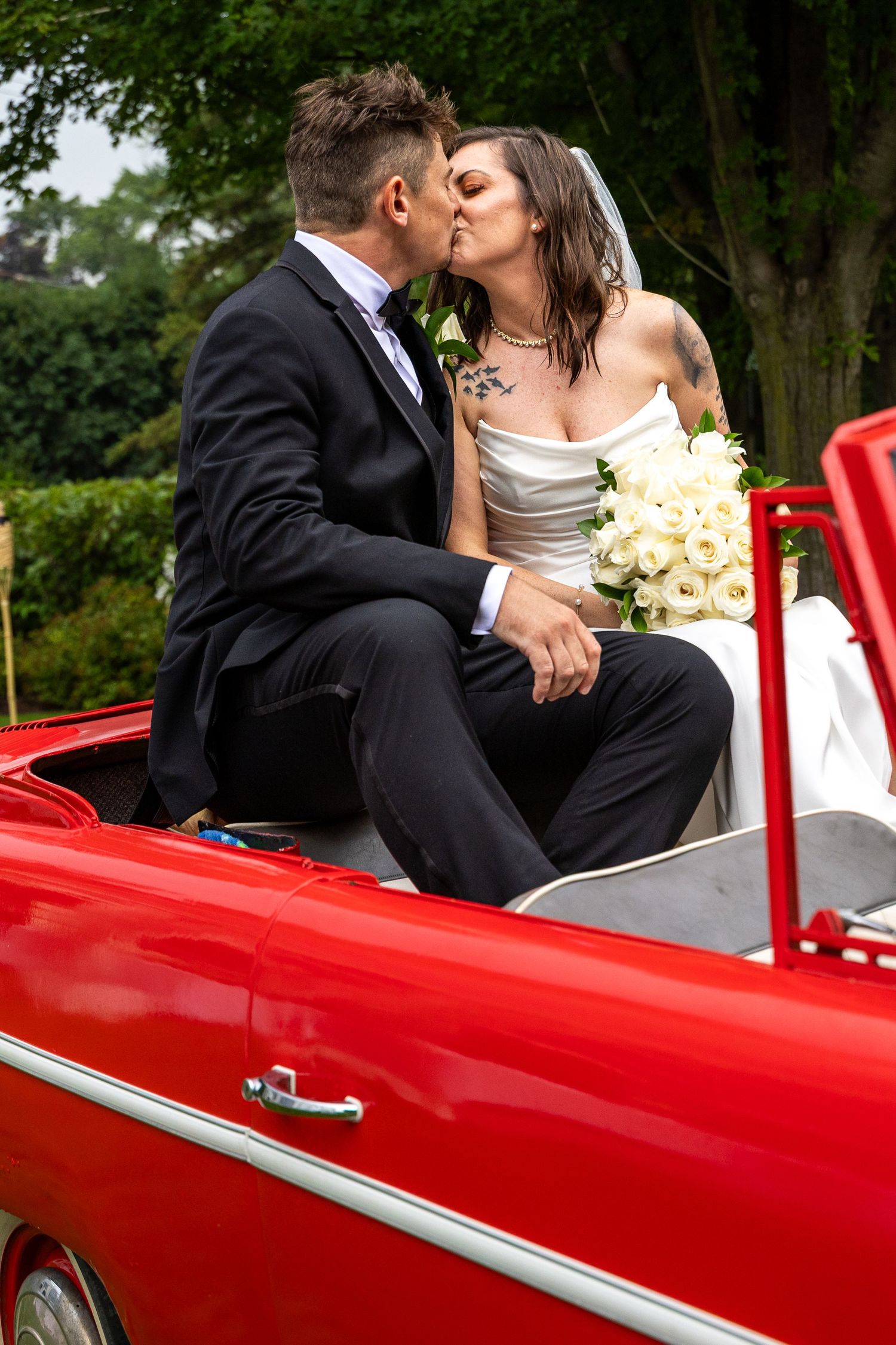 Newlywed couple sharing a passionate kiss seated on a classic red convertible car outdoors.