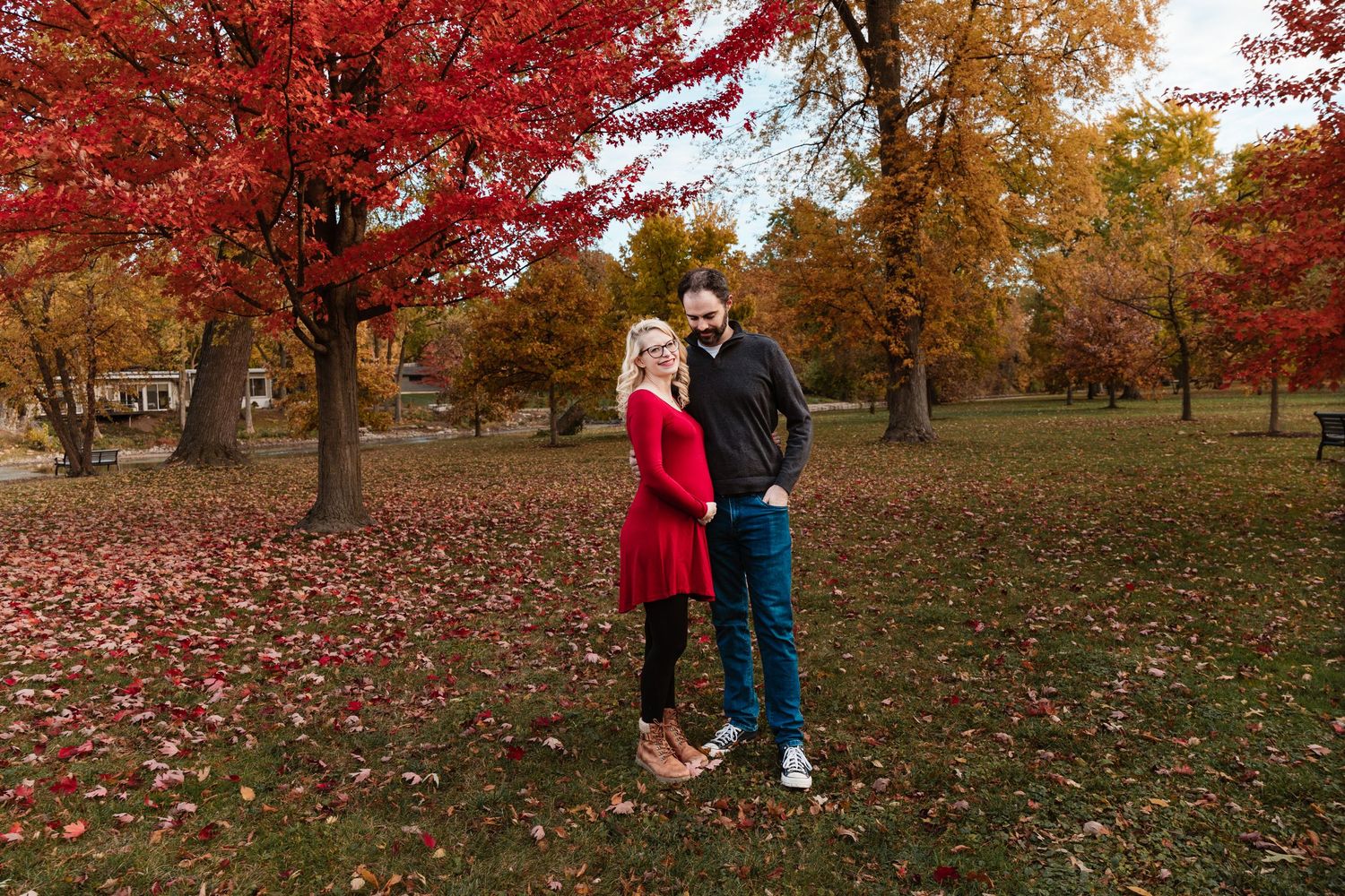 Couple holding hands in red autumn park for maternity photo session among colorful fall foliage.