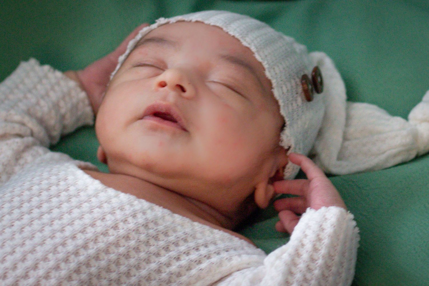 Peaceful newborn baby in white knit hat sleeping on soft green background.