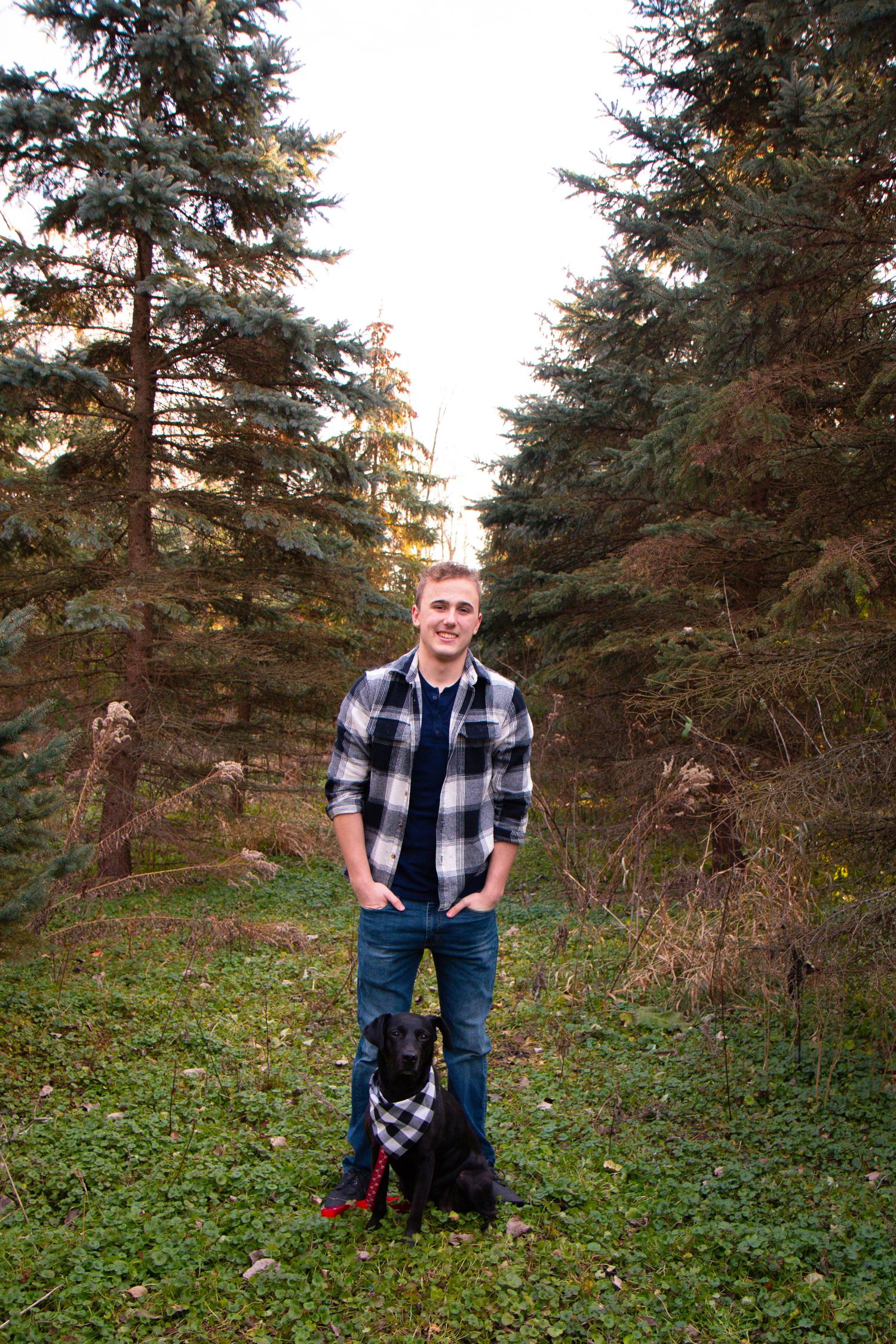 Young man in plaid shirt smiling with black dog in forest clearing surrounded by evergreen trees.