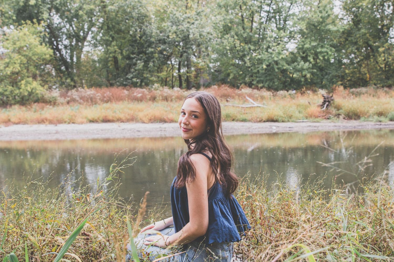Young woman with long brown hair sitting by a serene pond surrounded by autumn foliage and tall grass.