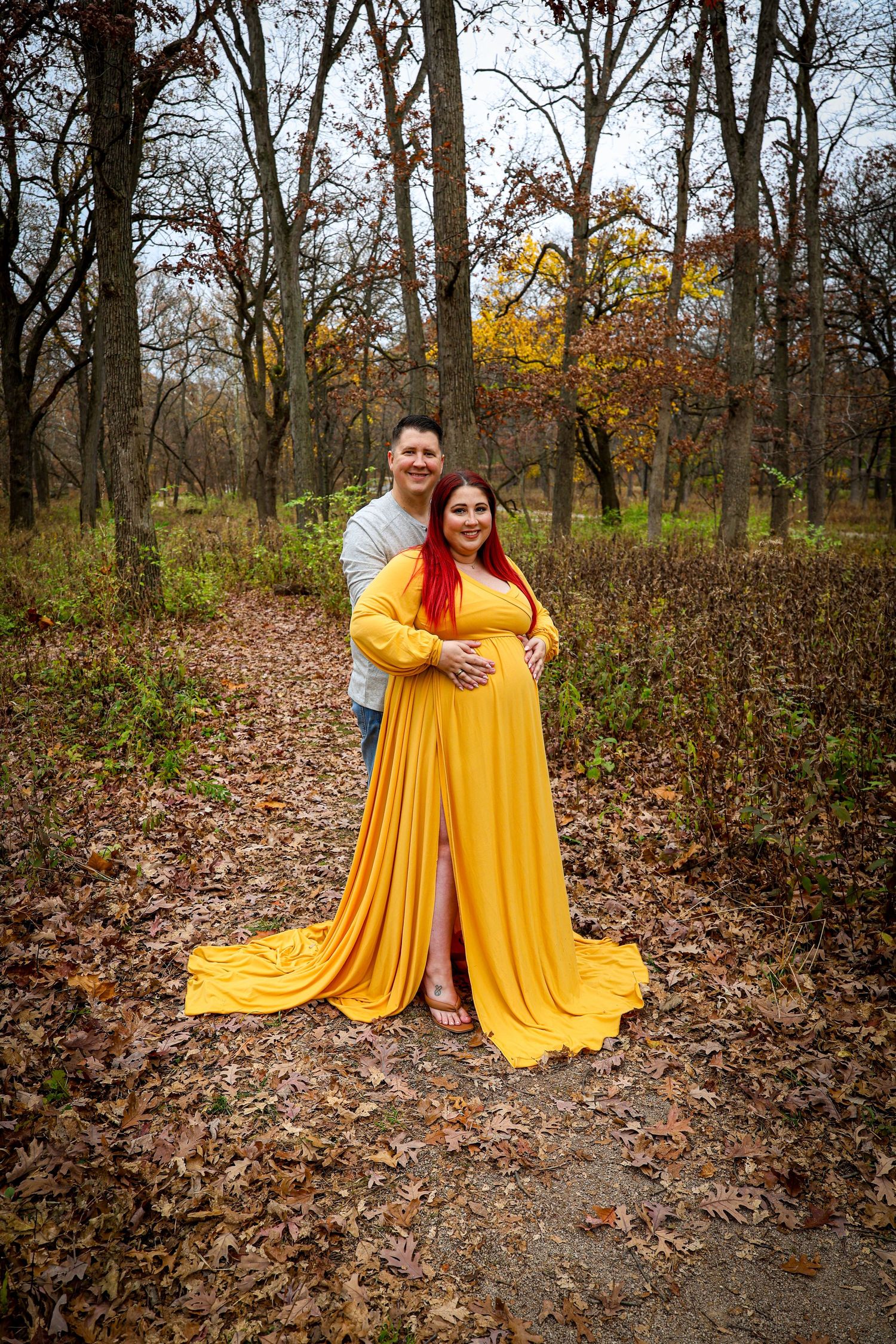 Couple maternity photo with woman in flowing yellow gown and man in autumn forest setting.