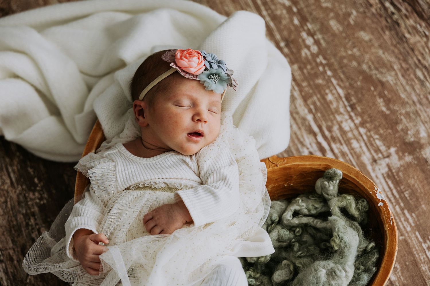 Sleeping newborn baby girl in white lace gown with floral headband resting in a wooden bowl.