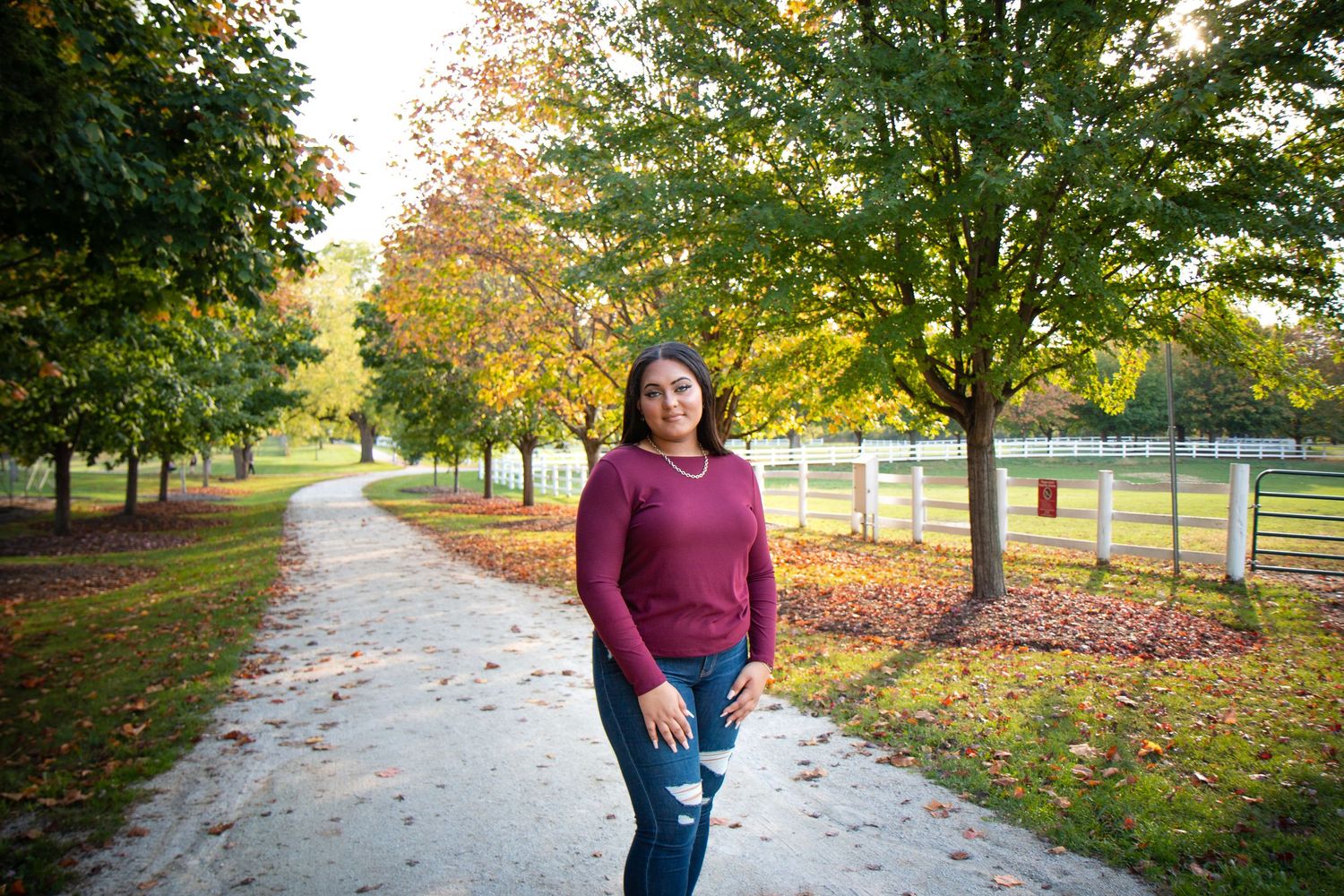 Young woman in burgundy sweater and jeans poses on a tree-lined path in autumn park with colorful foliage.