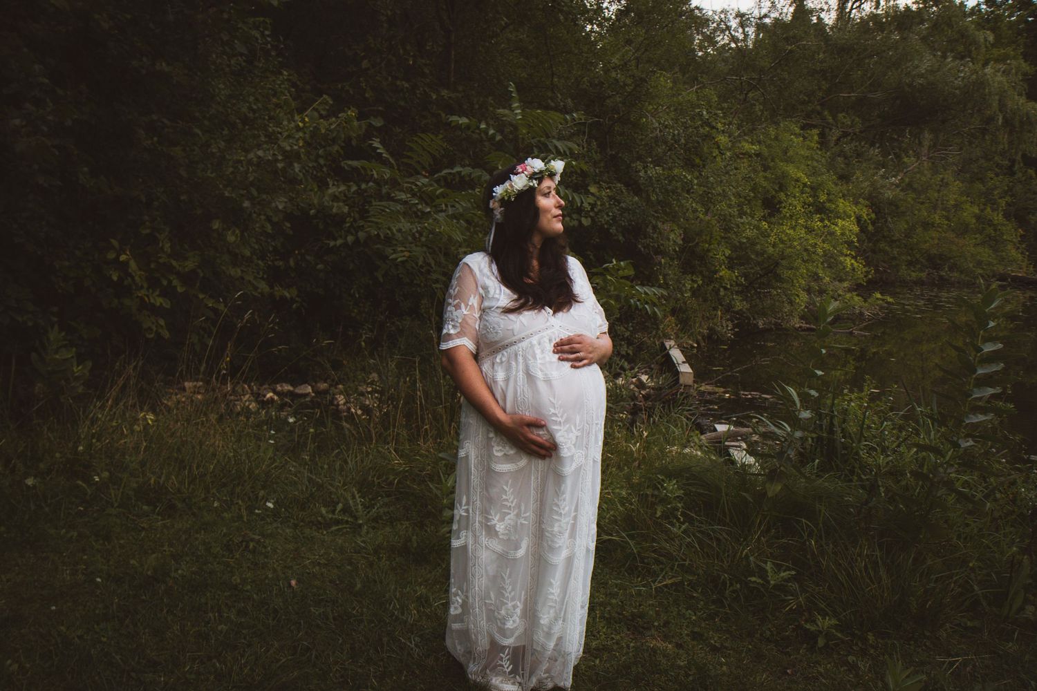 Pregnant woman in white lace dress and floral crown standing in lush green garden setting.