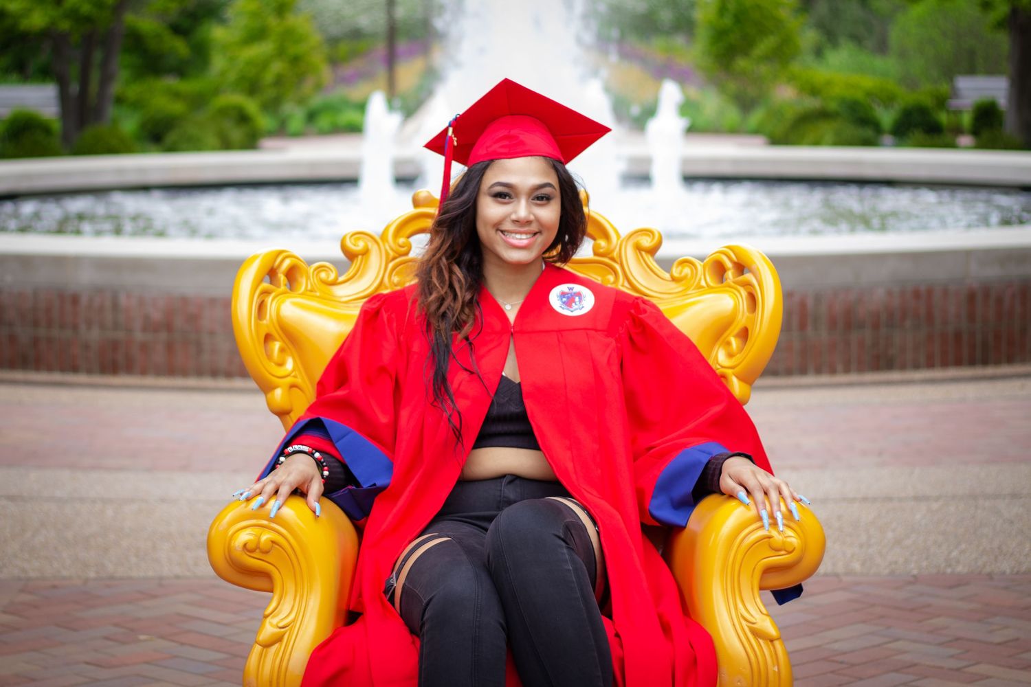 Graduate in red cap and gown sits on golden throne chair outdoors near fountain for graduation portrait.