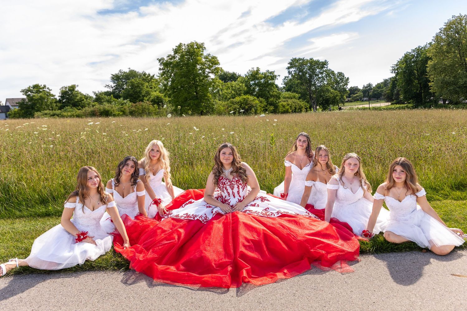 Quinceañera girl in red and white gown poses with chambelanes in white dresses spread across a sunny outdoor field.