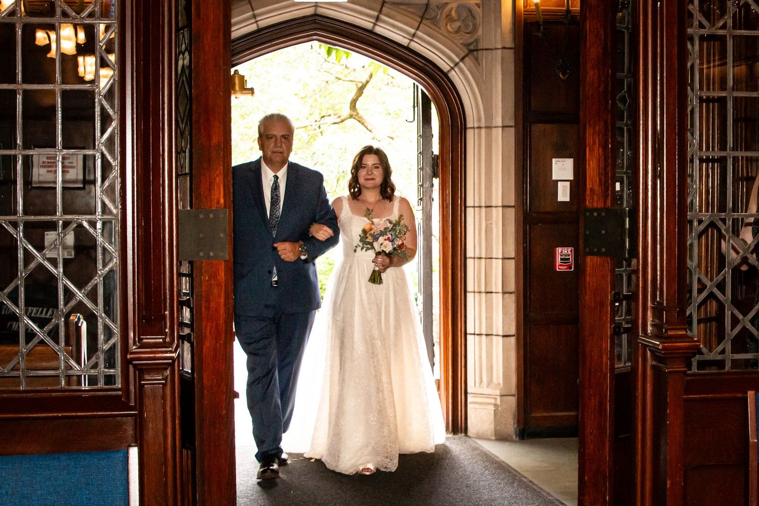 A bride in a flowing gown is escorted through ornate church doors by her father on her wedding day.