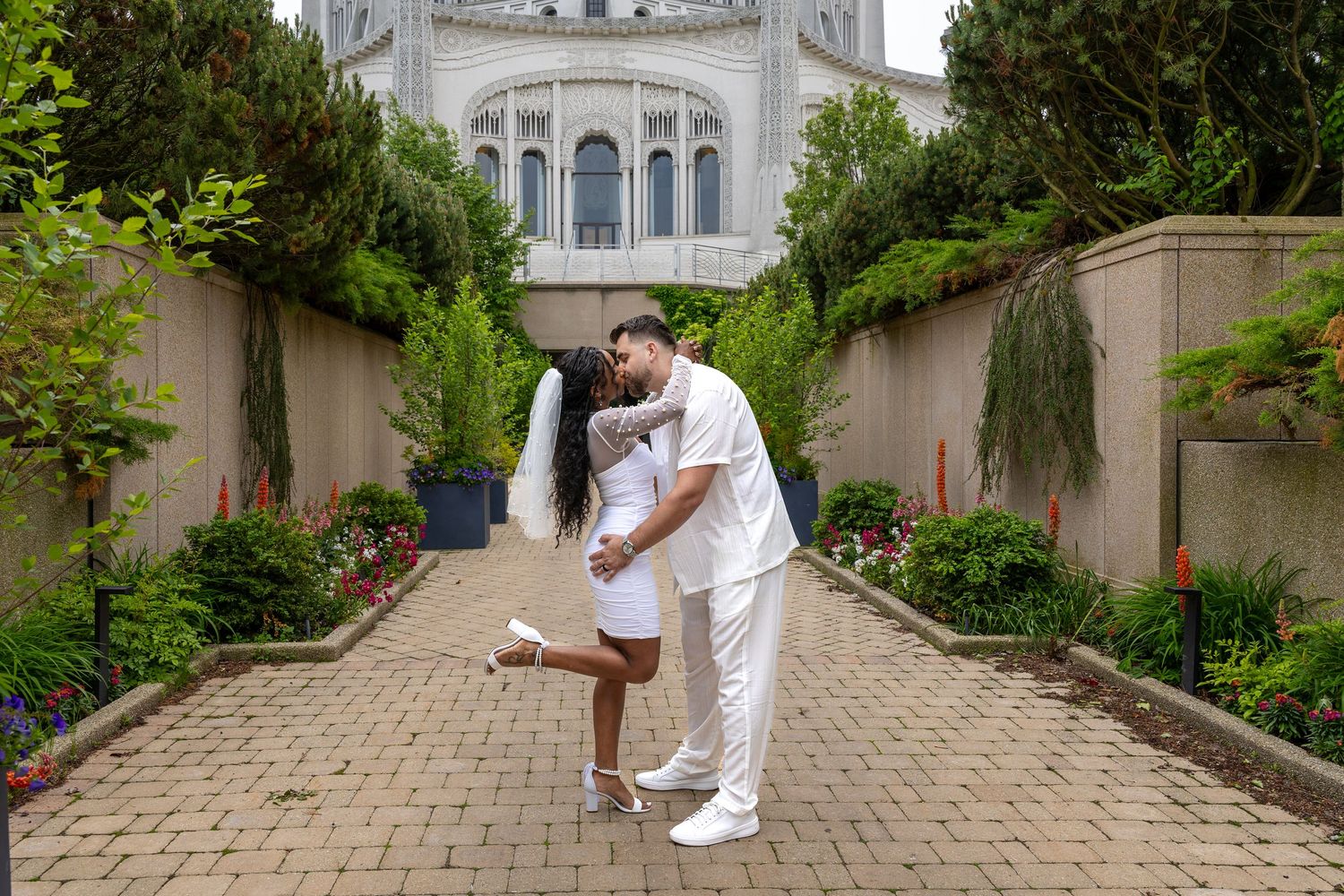 A couple shares a romantic dip kiss on a brick pathway surrounded by lush gardens and an elegant building.