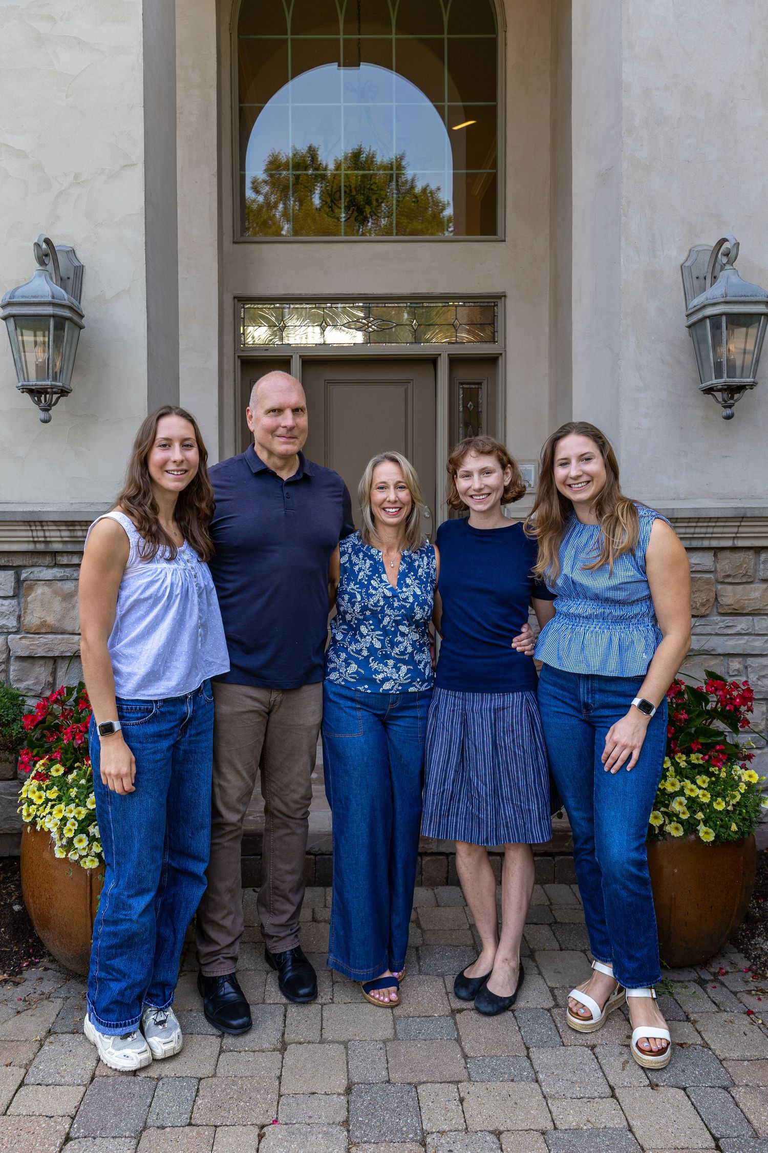 A family of five smiling together in front of an elegant home entrance with flower pots and stone facade.