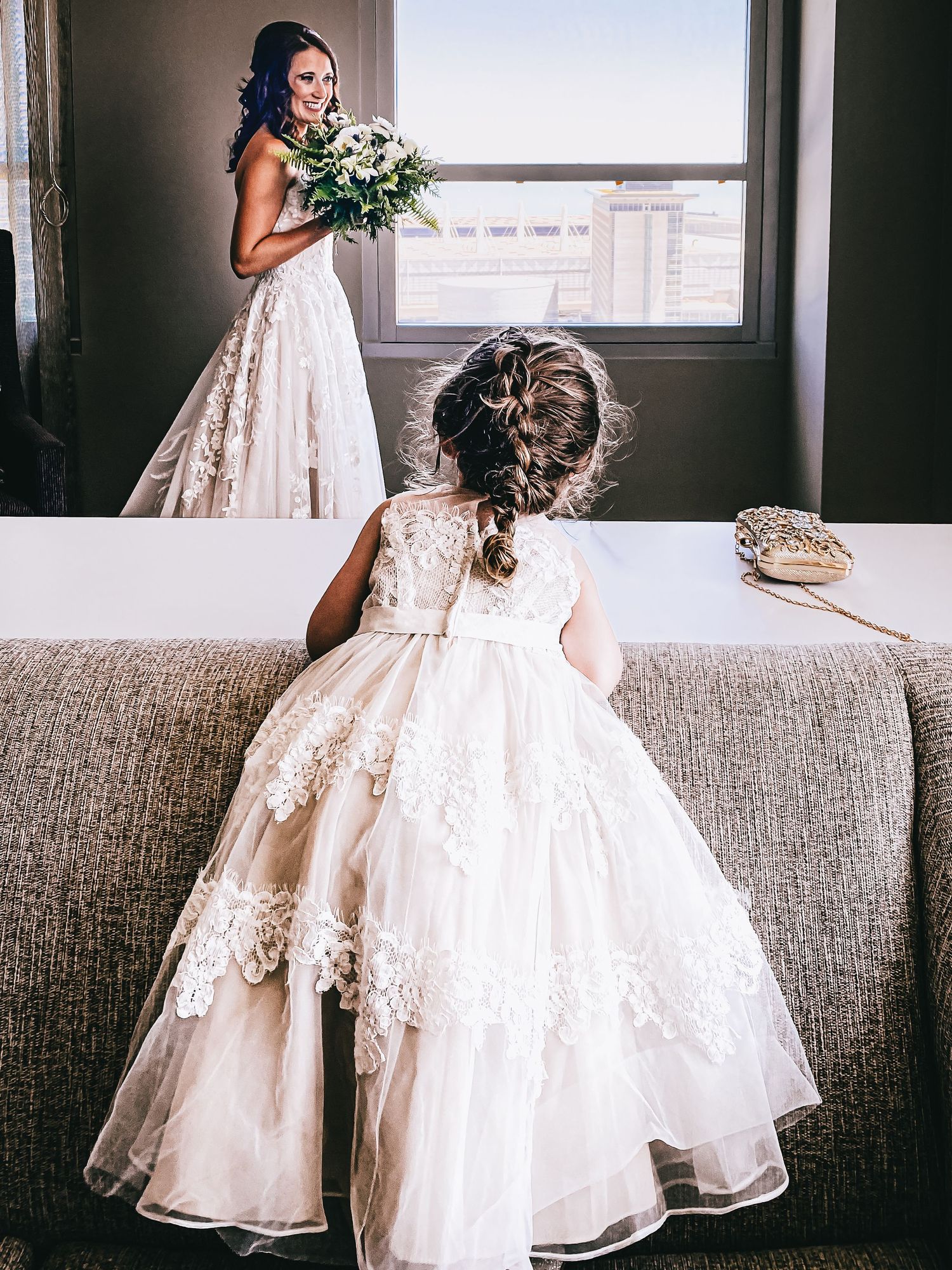 Bride holding floral bouquet stands by window as flower girl in lace dress watches from sofa.