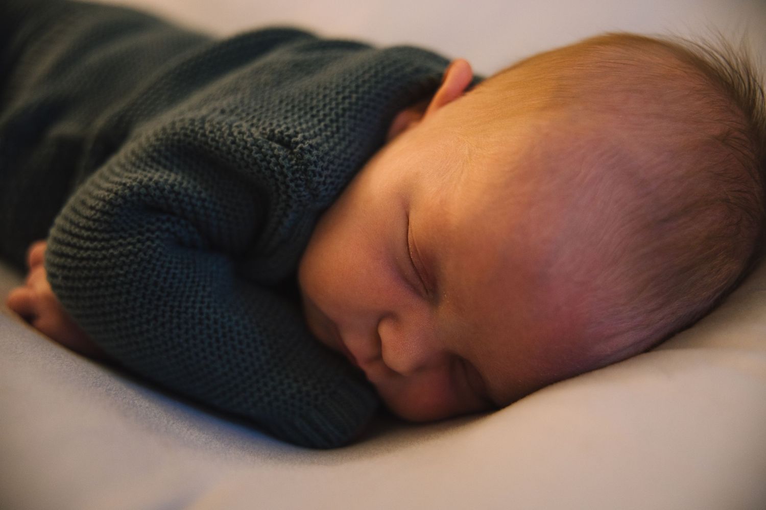 Peaceful newborn baby sleeping soundly in a dark teal knit outfit on a soft white surface.