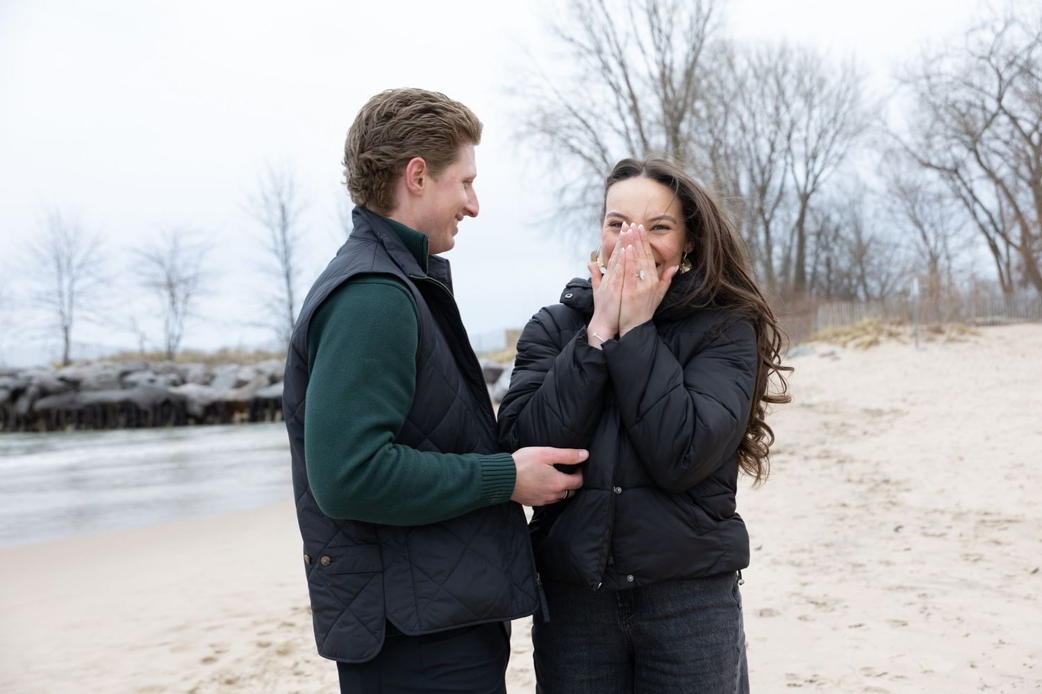 Couple sharing a romantic moment on a sandy beach, woman covering her mouth in surprise while man smiles beside her.