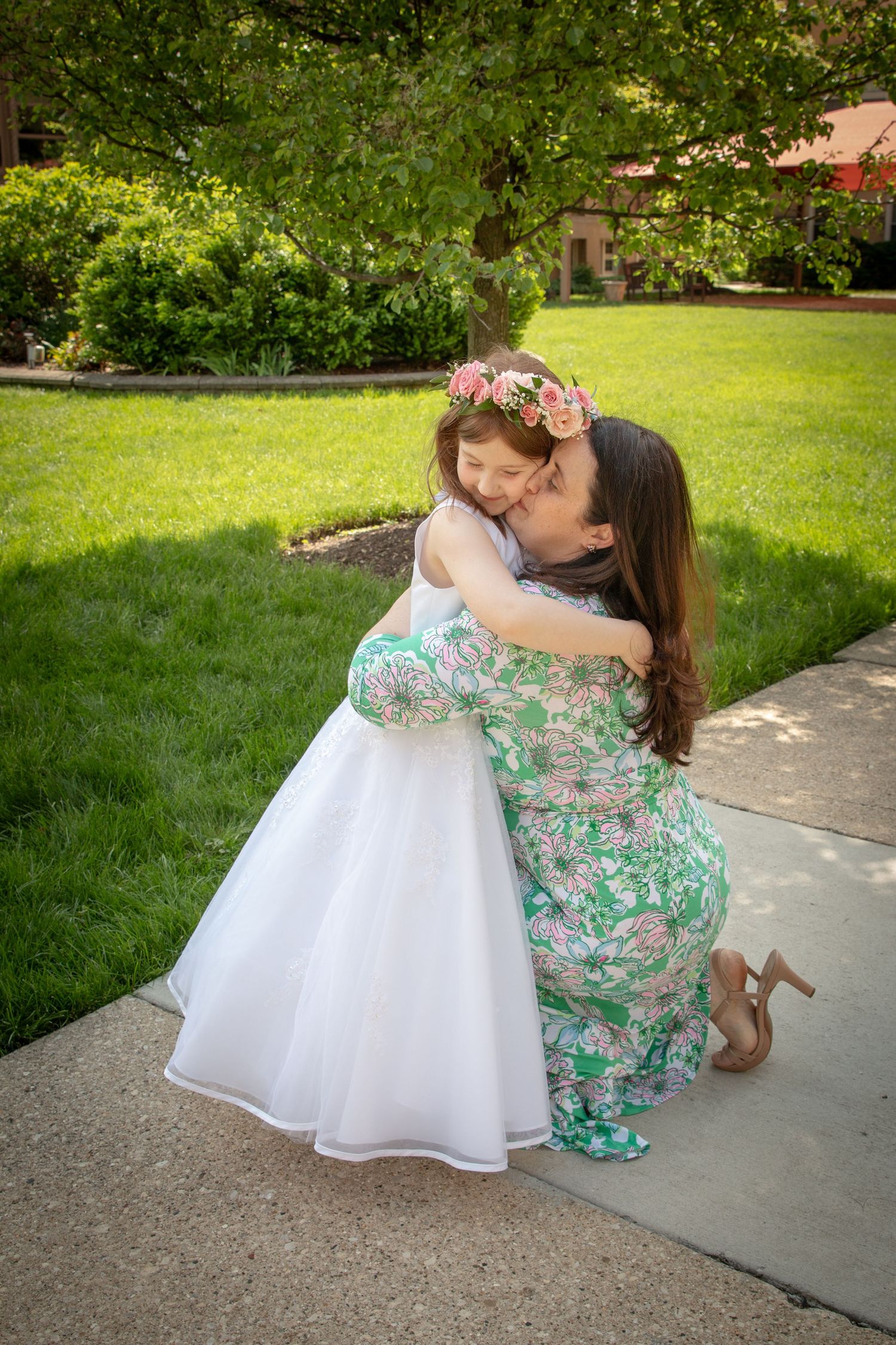 Woman in floral dress kneels to hug young girl in white dress with flower crown outdoors.