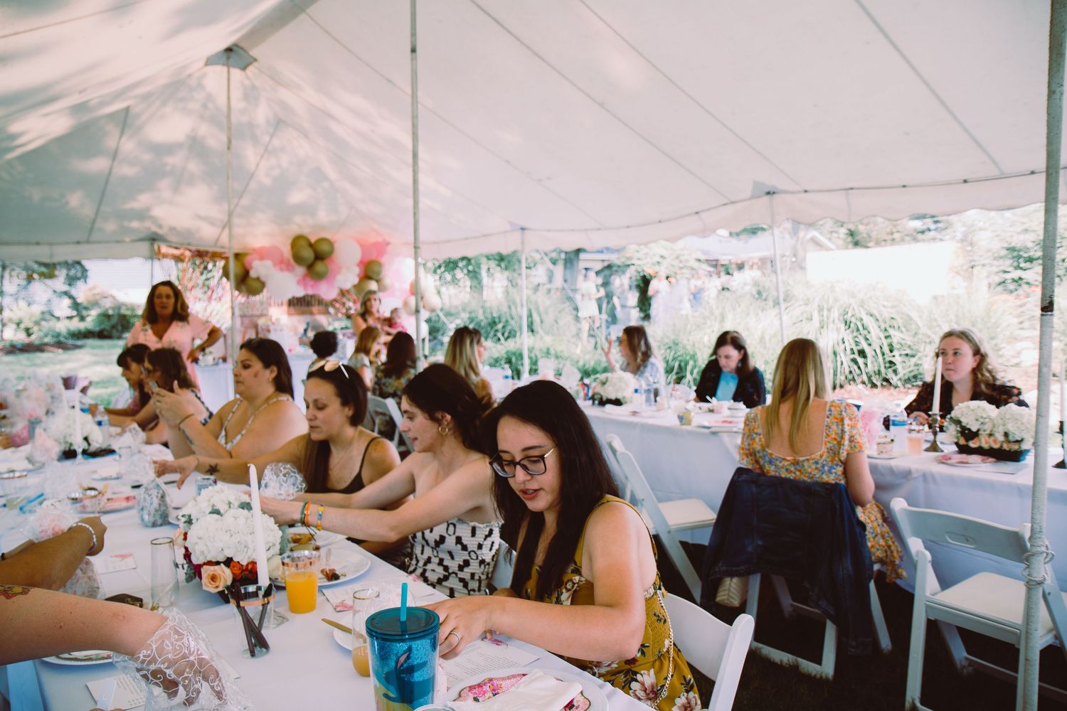 Women guests seated at long tables under a white tent at an outdoor bridal shower celebration.