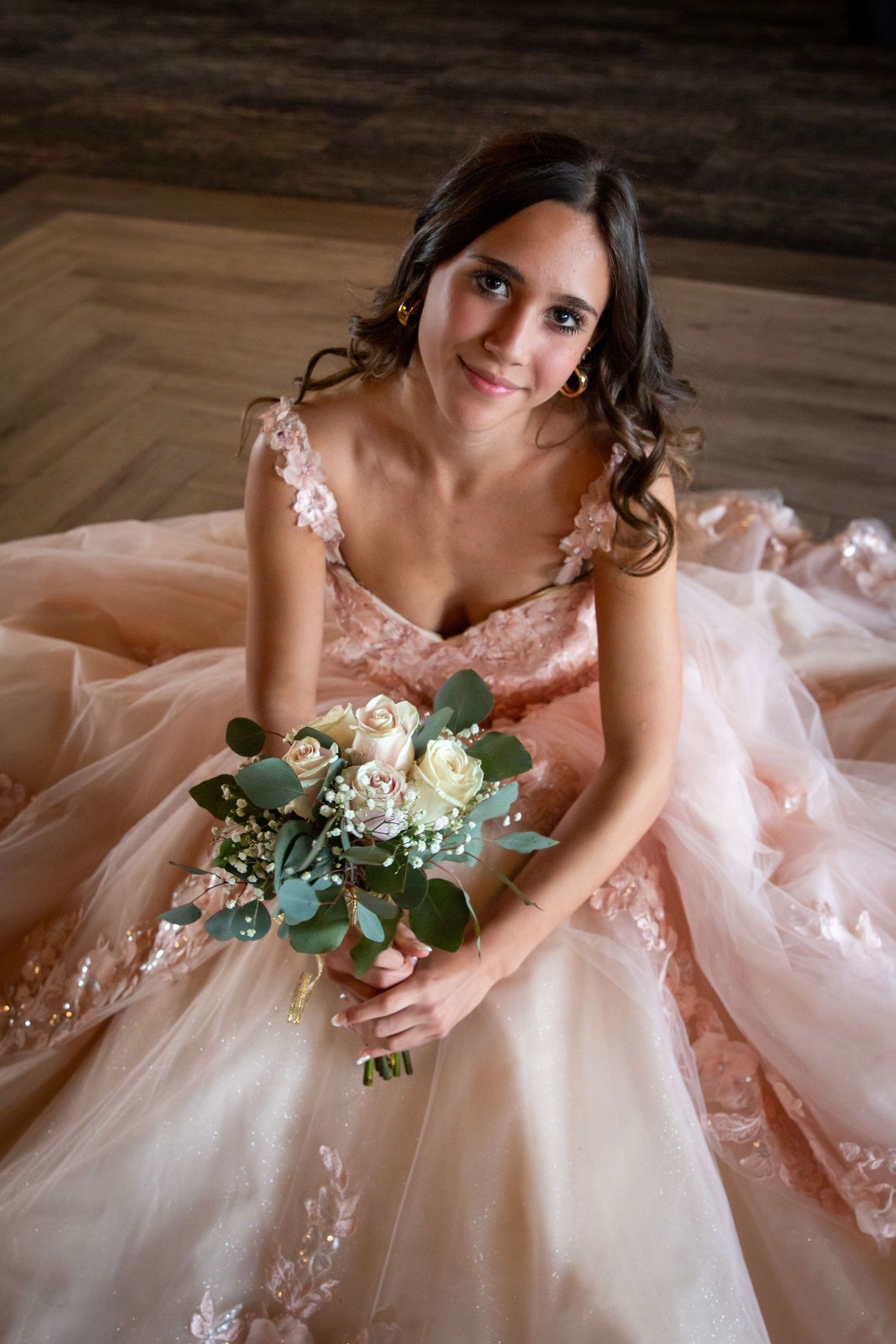 Young bride in blush pink ballgown sits on floor holding white rose bouquet, smiling up at camera.
