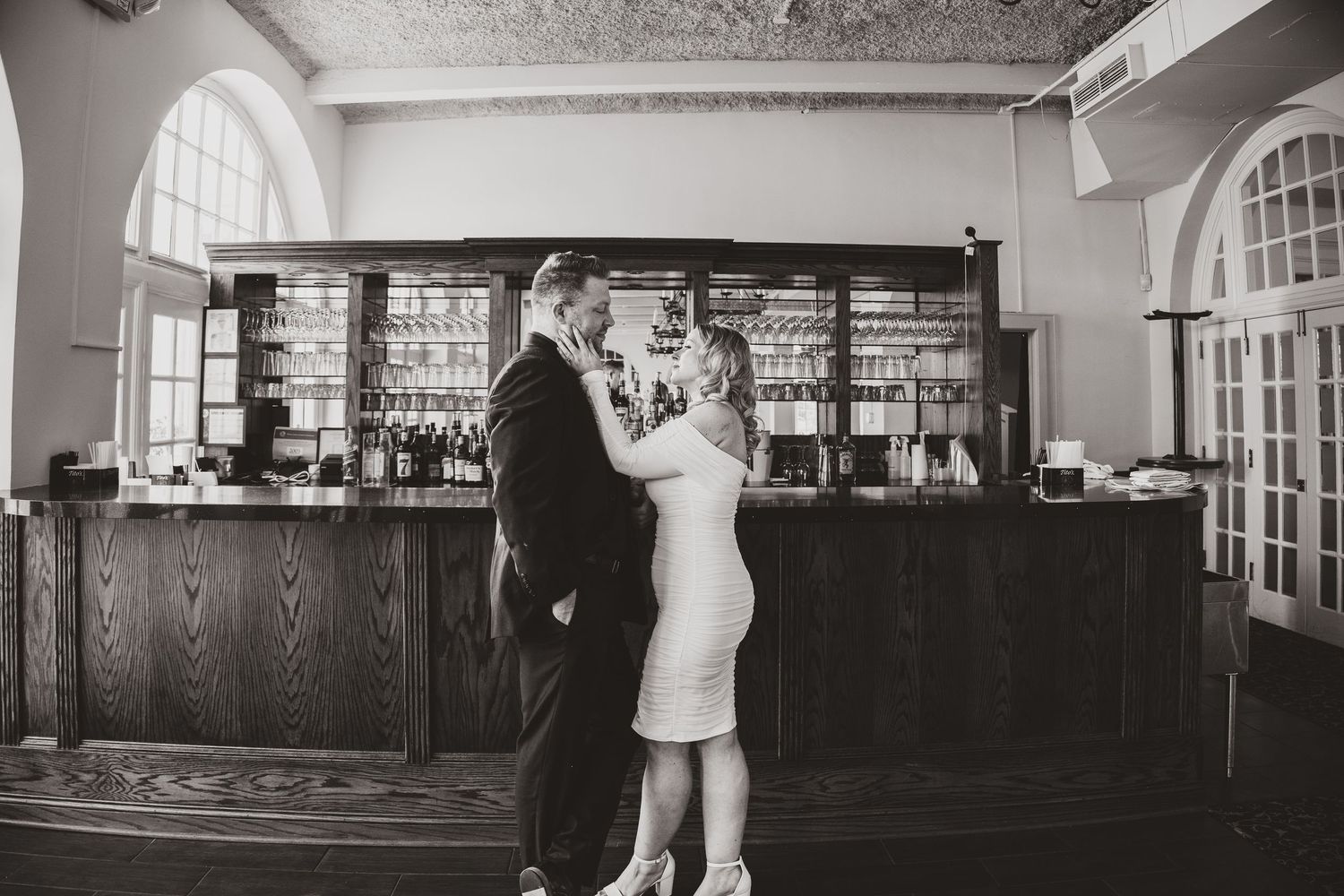 Black and white photo of a couple embracing and dancing in front of an elegant bar with shelves of bottles.