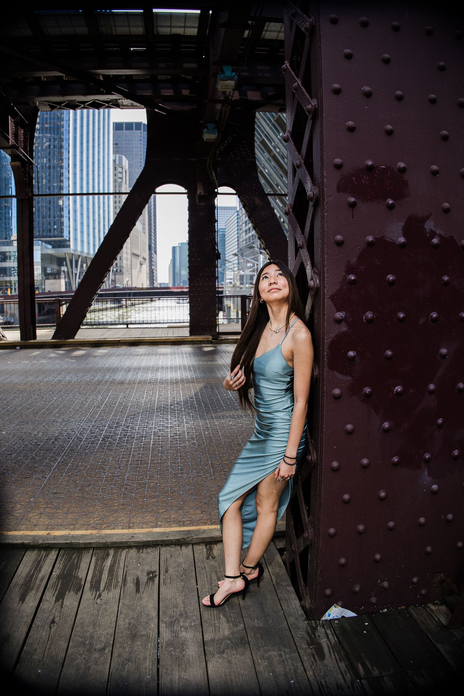 Woman in teal dress poses against rustic steel bridge structure with Chicago city skyline in background.