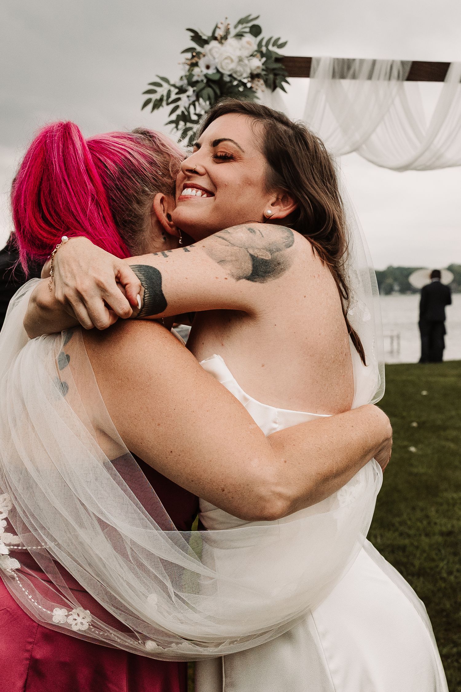 Two brides share a heartfelt embrace near a floral arch during an outdoor lakeside wedding ceremony.