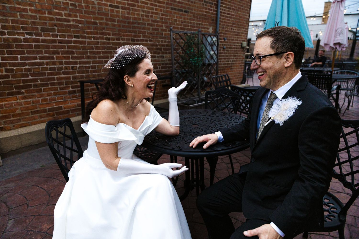 A laughing bride in white gloves and groom share a joyful moment seated at an outdoor brick patio.