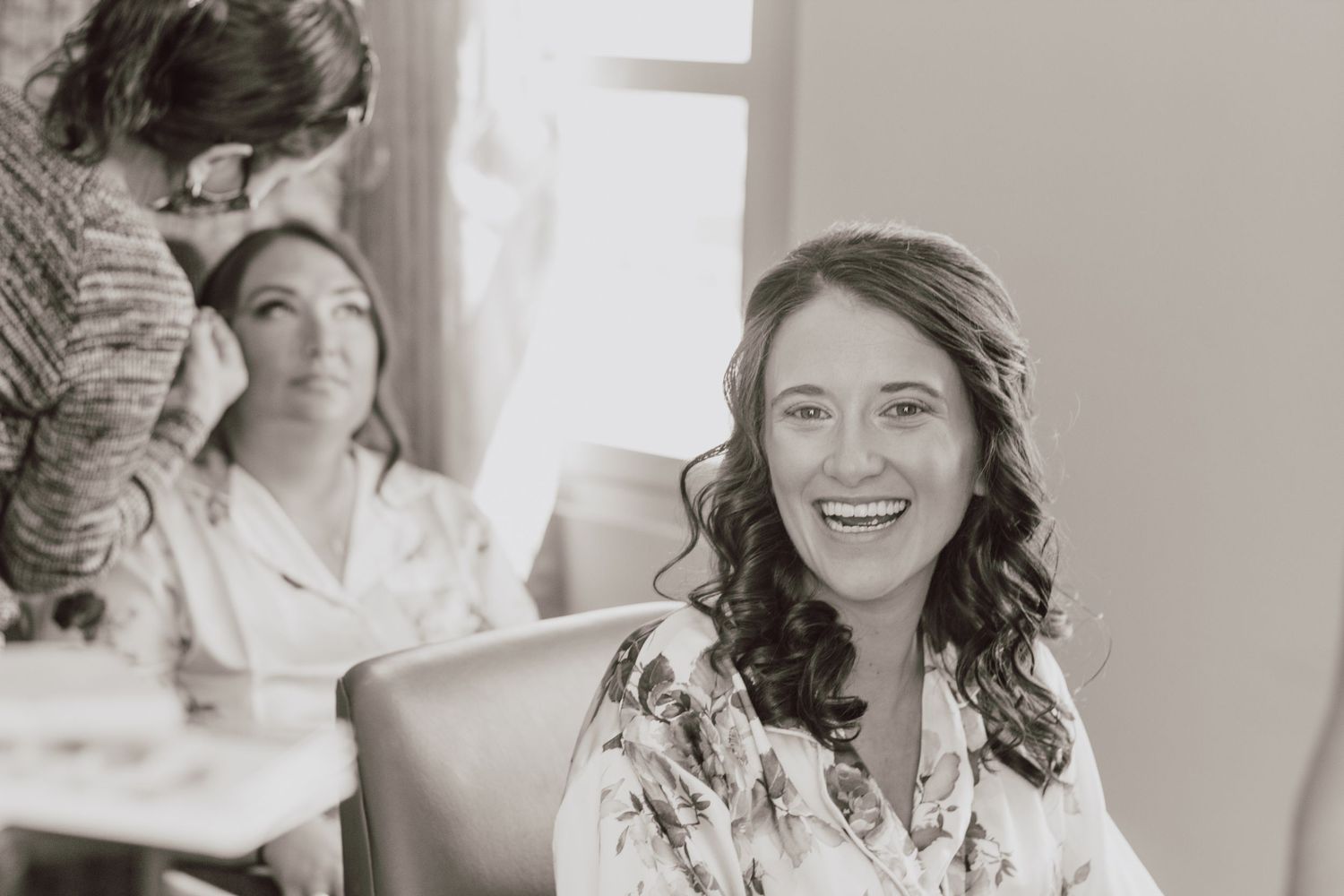 Smiling bride in floral robe laughs joyfully during hair and makeup preparations on wedding day.