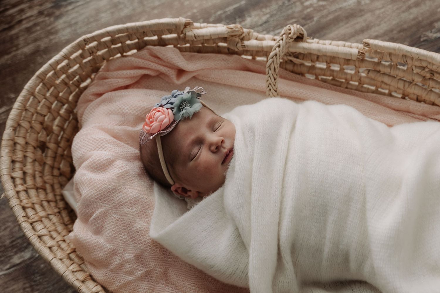 Newborn baby girl in white swaddle with floral headband sleeping in a wicker Moses basket.