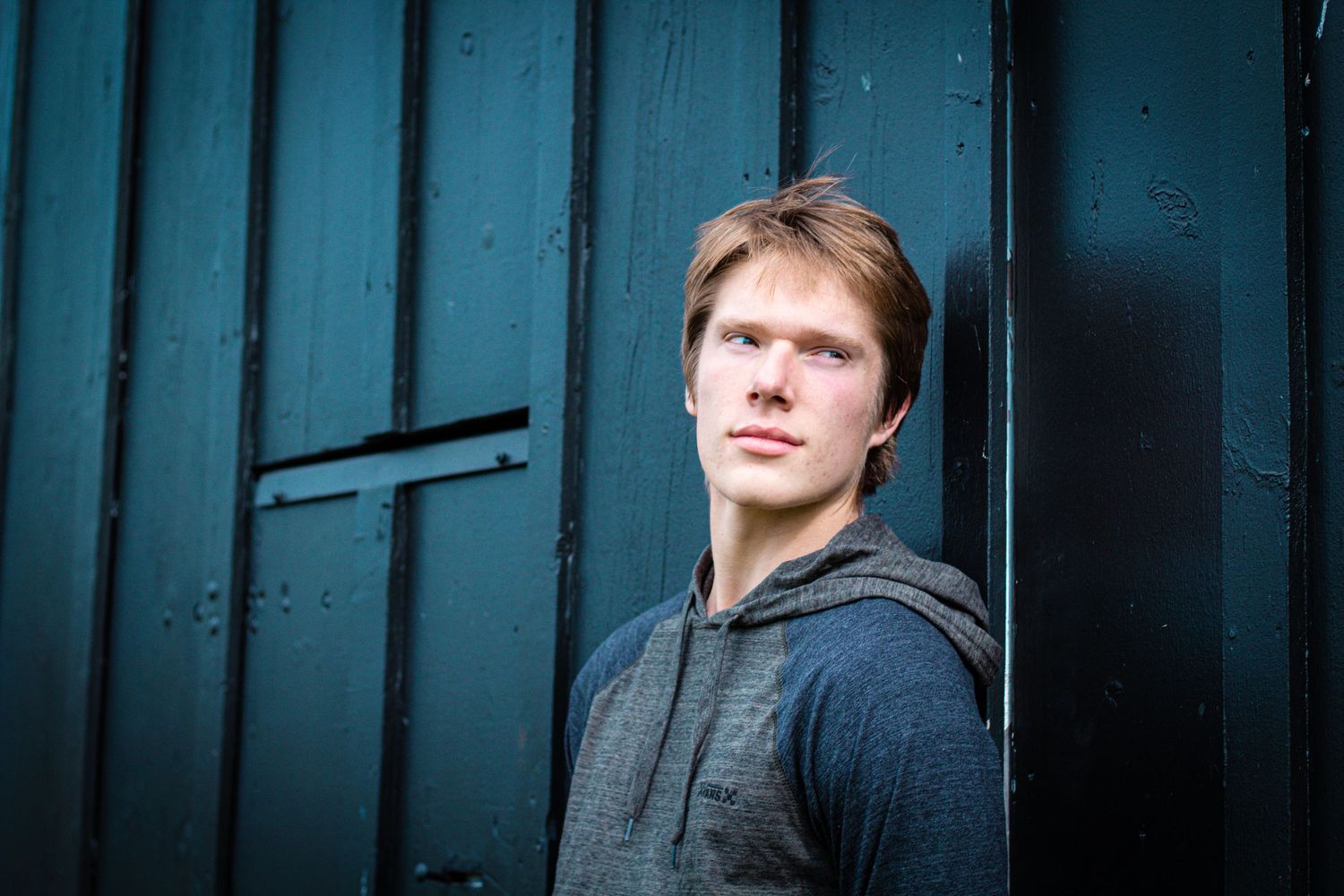 Young man with blonde hair leaning against a teal wooden door wearing a gray hoodie.