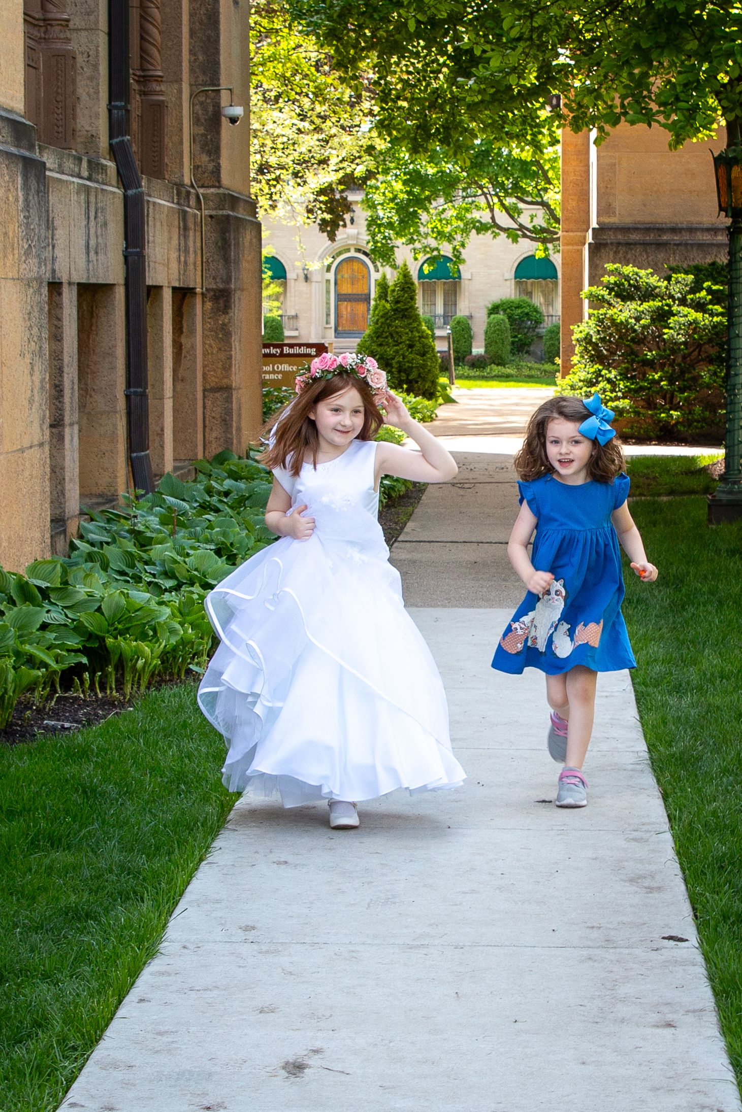 Two young girls in dresses run along sidewalk outside stone building on sunny day.