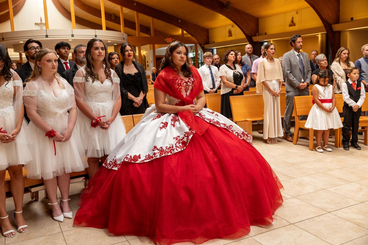 Young woman in red and white quinceañera gown stands during a church ceremony with guests in white.