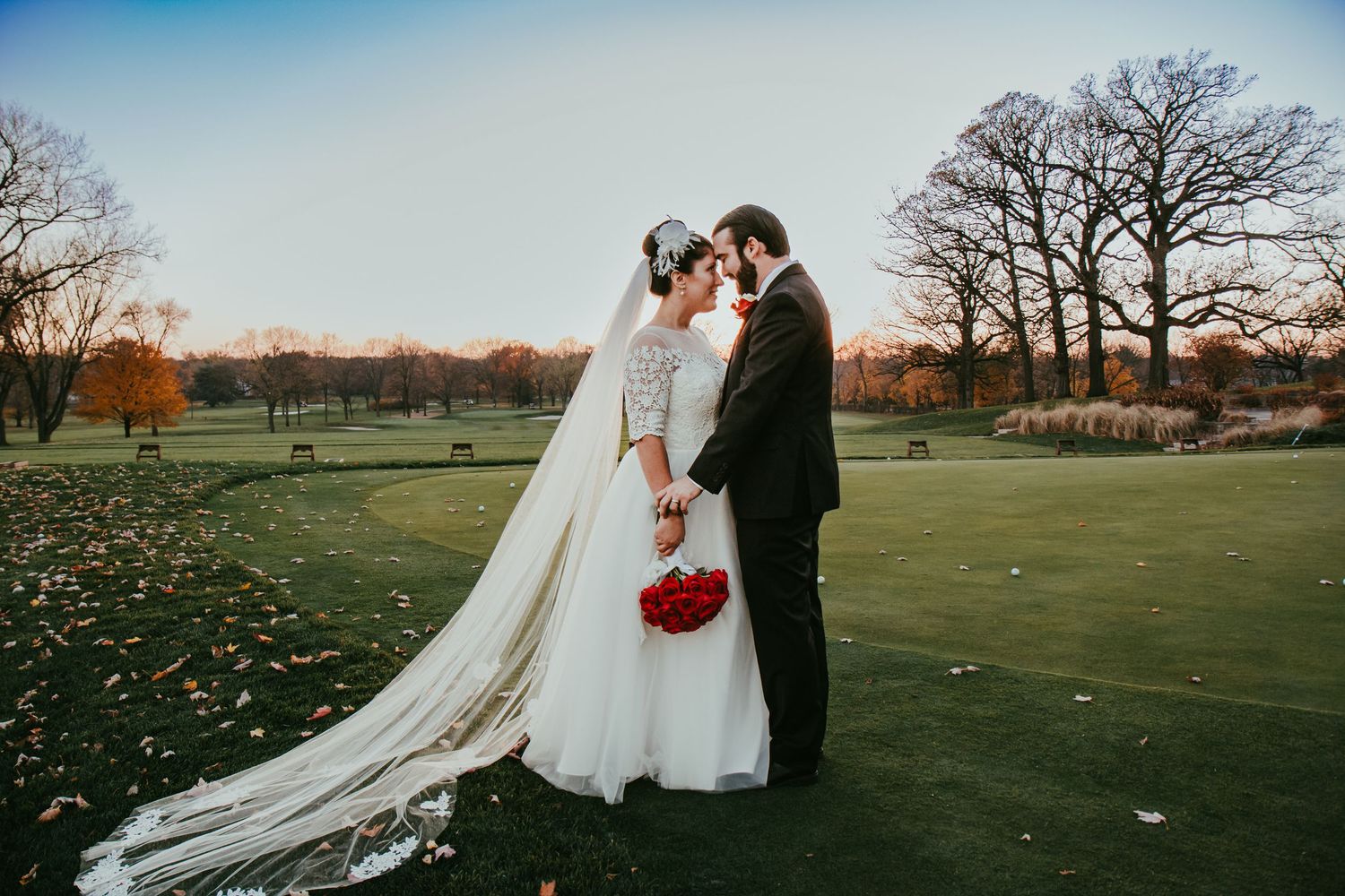 Bride and groom touch foreheads tenderly on golf course with autumn trees at golden hour.
