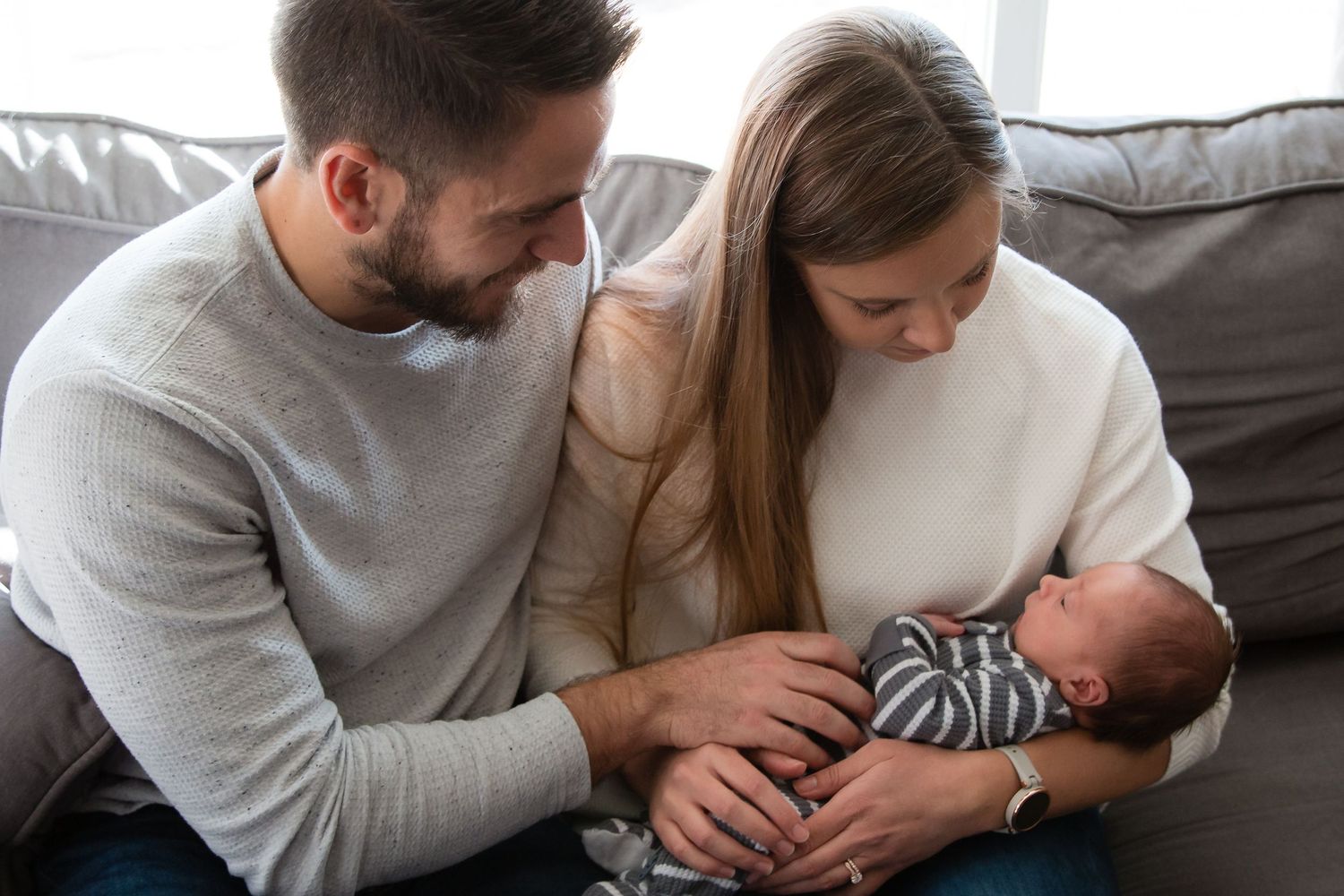 New parents sitting on a gray couch admiring their sleeping newborn baby together.