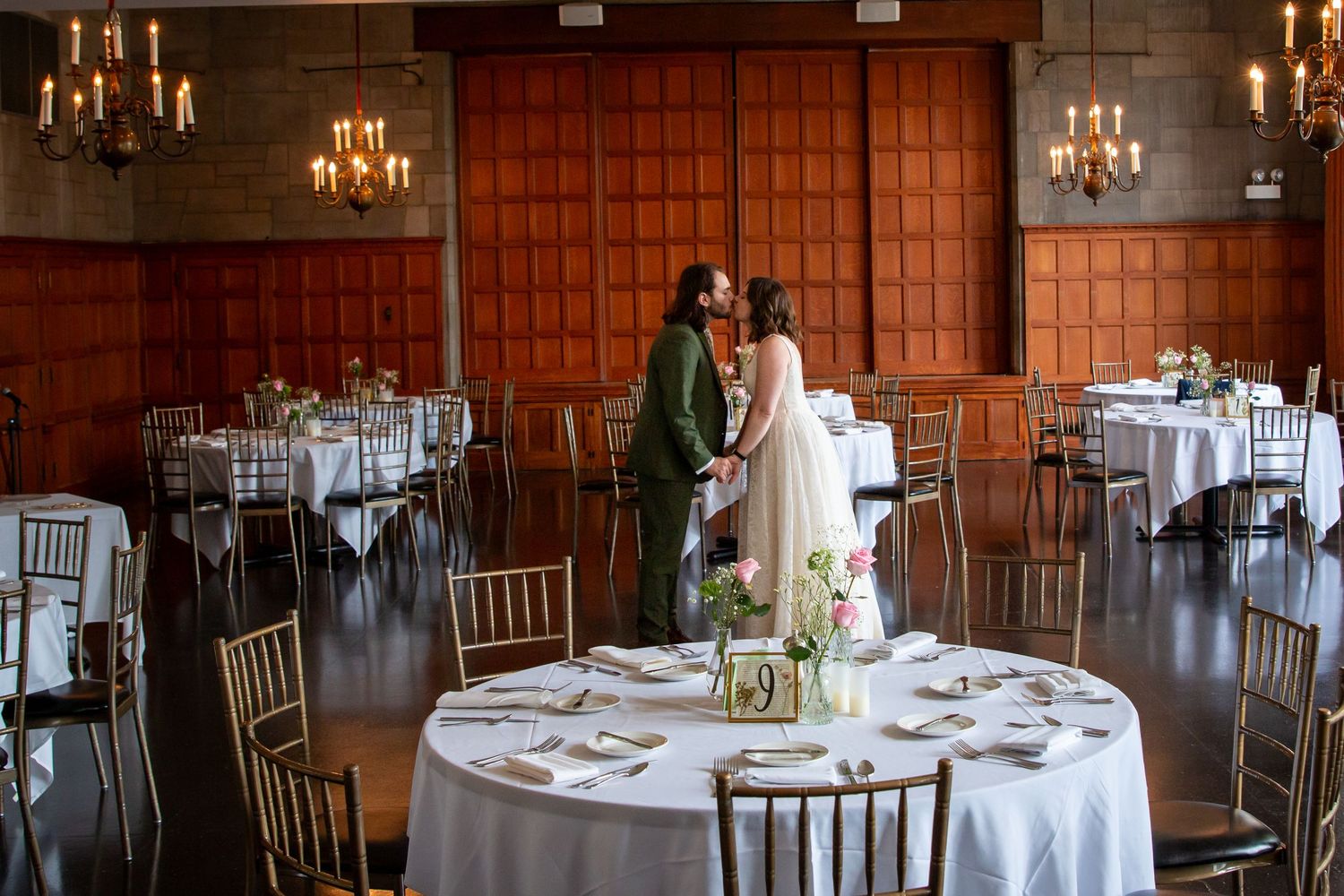 Bride and groom share a kiss in an elegant wedding reception hall with candlelit chandeliers and round tables.