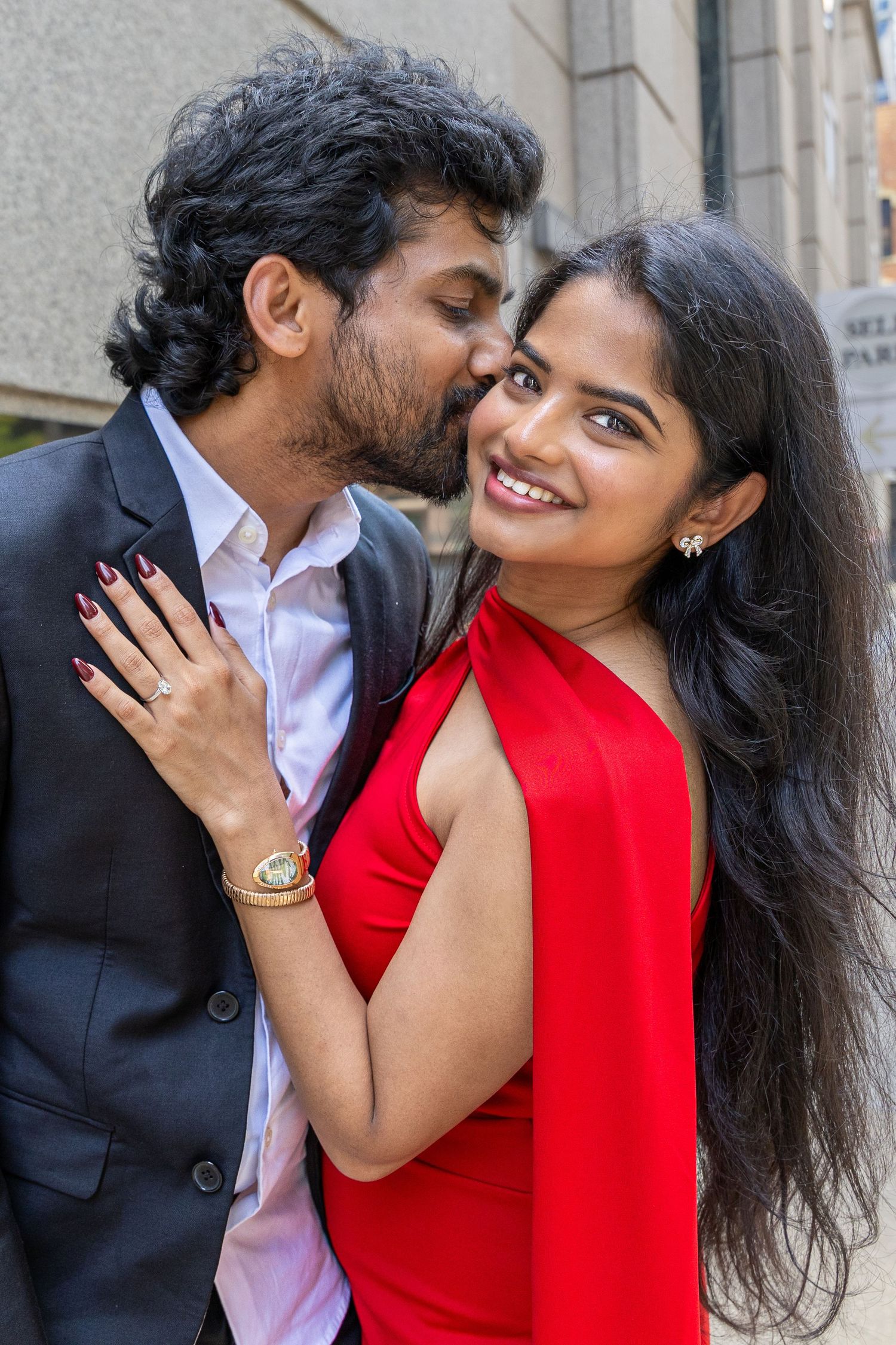 A couple in formal wear shares a romantic moment, man in suit kisses woman in red dress who smiles joyfully.