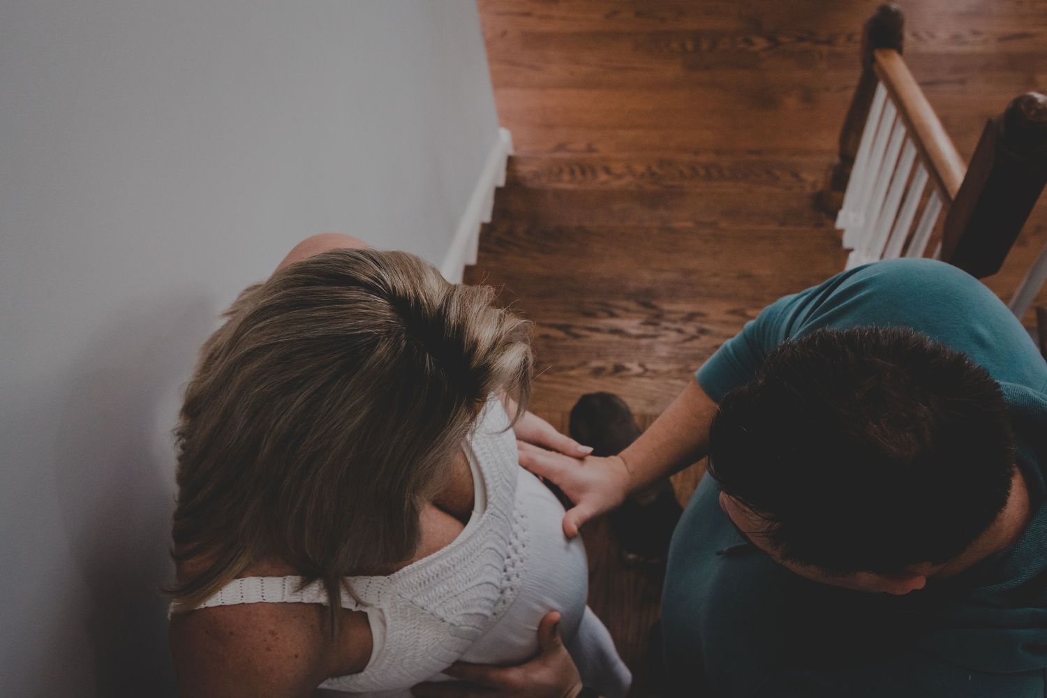 Overhead view of two adults lovingly cradling a baby on wooden stairs indoors.
